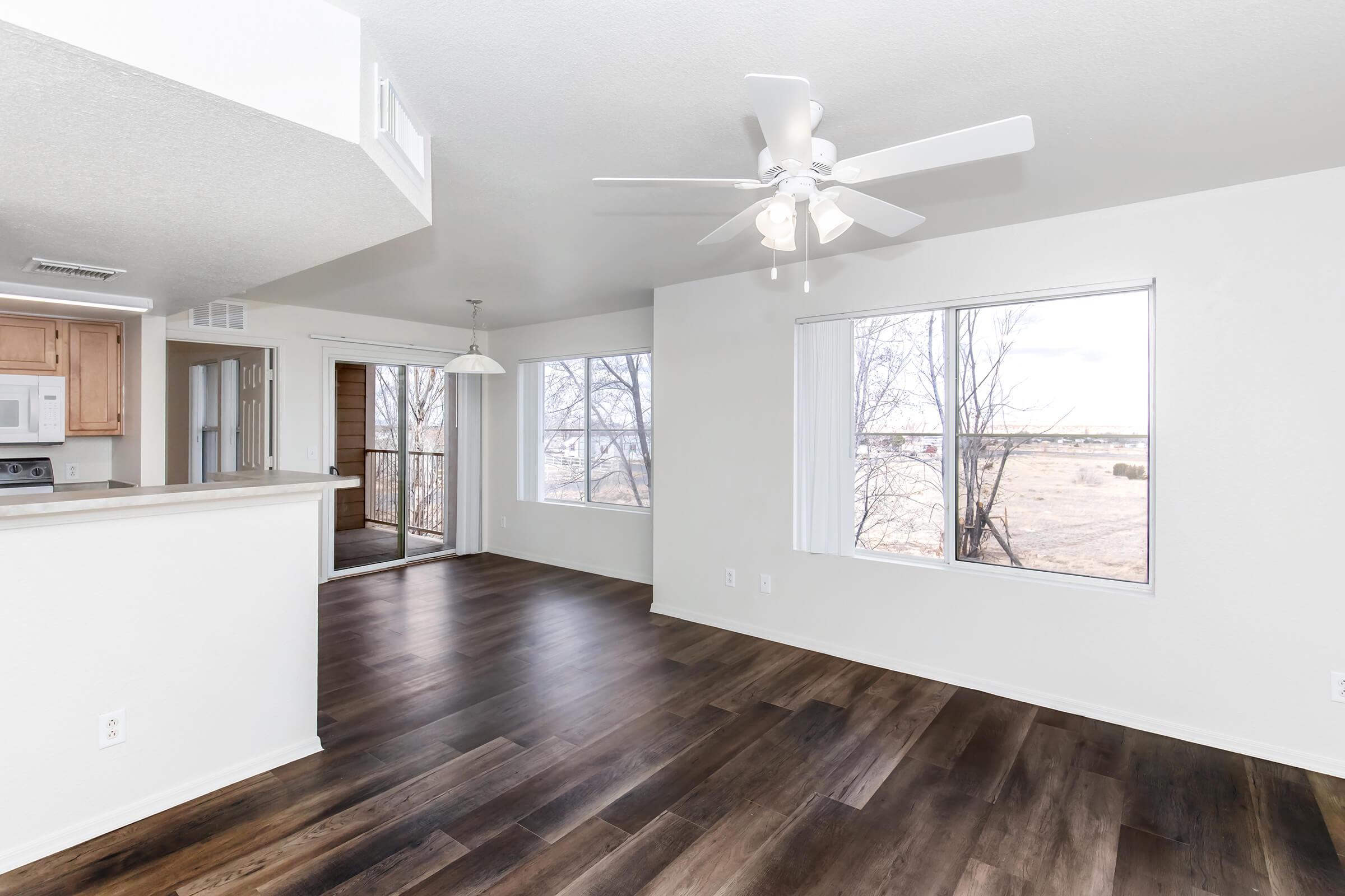 Bright, spacious living area with large windows allowing natural light. Features a ceiling fan and open layout connecting to a kitchen. Light-colored walls and dark wood flooring create a modern ambiance. A sliding door leads to a patio area with views of a natural landscape.