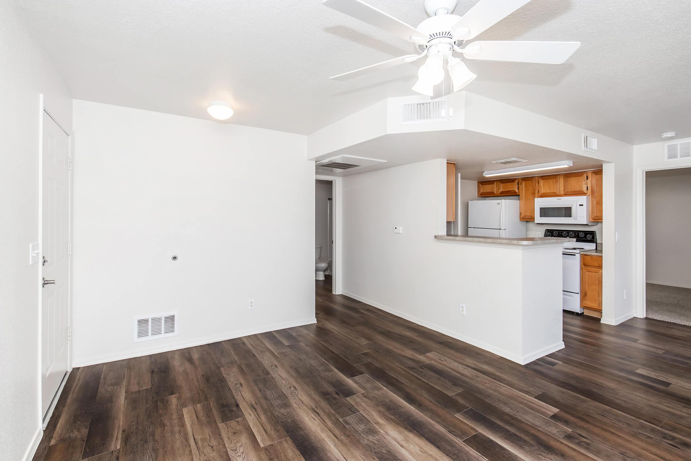 A spacious interior of an apartment featuring a living area with wood-like flooring, a ceiling fan, and a kitchen with wooden cabinets and appliances. There are doorways leading to additional rooms, including a bathroom, and light-colored walls enhance the bright atmosphere.