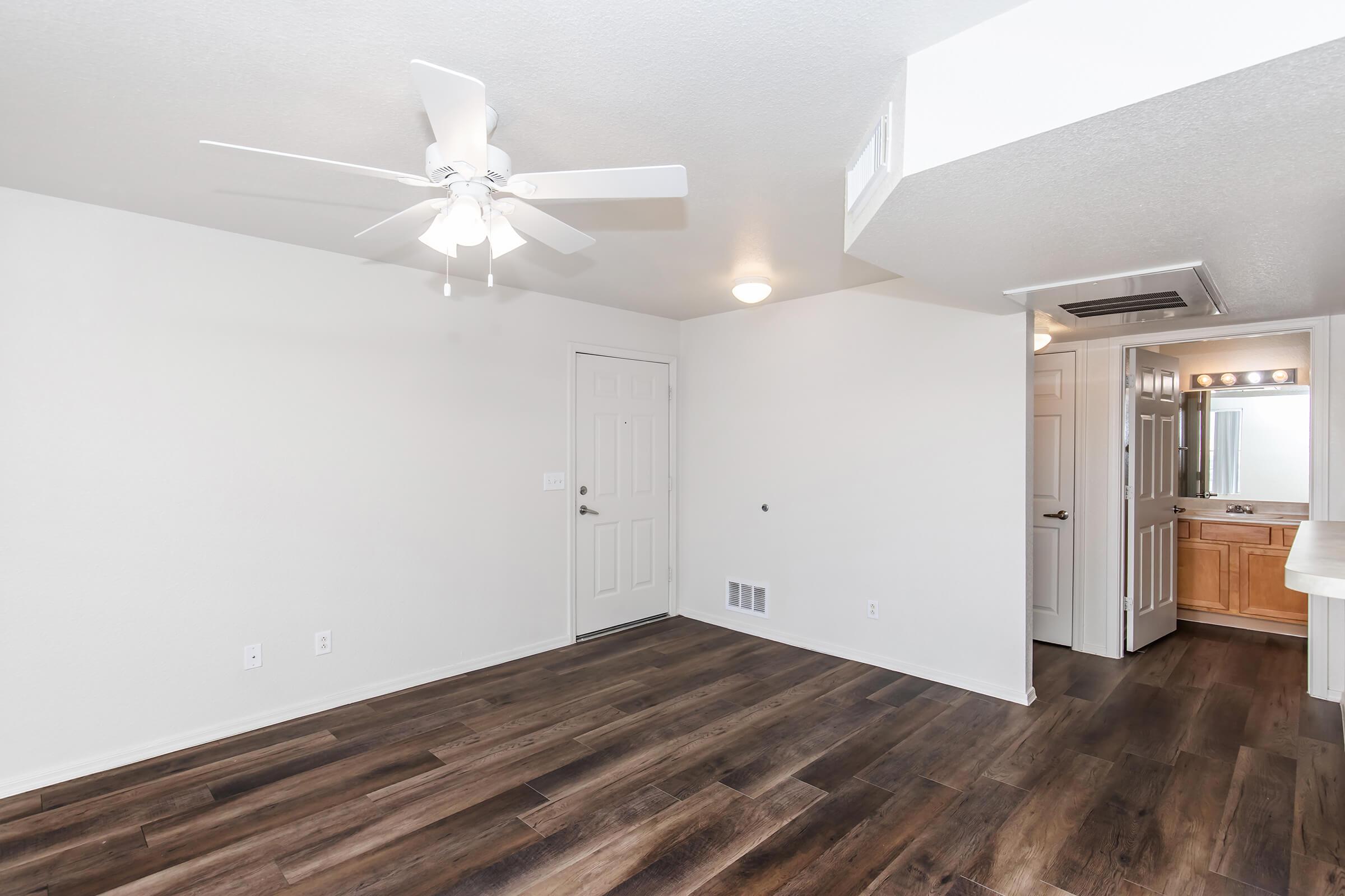 A bright, empty living space featuring a ceiling fan, light-colored walls, and dark wood flooring. A doorway leads to a bathroom with a lighted mirror, and there are no furnishings. The overall ambiance is clean and modern, with natural light coming from an unseen window.