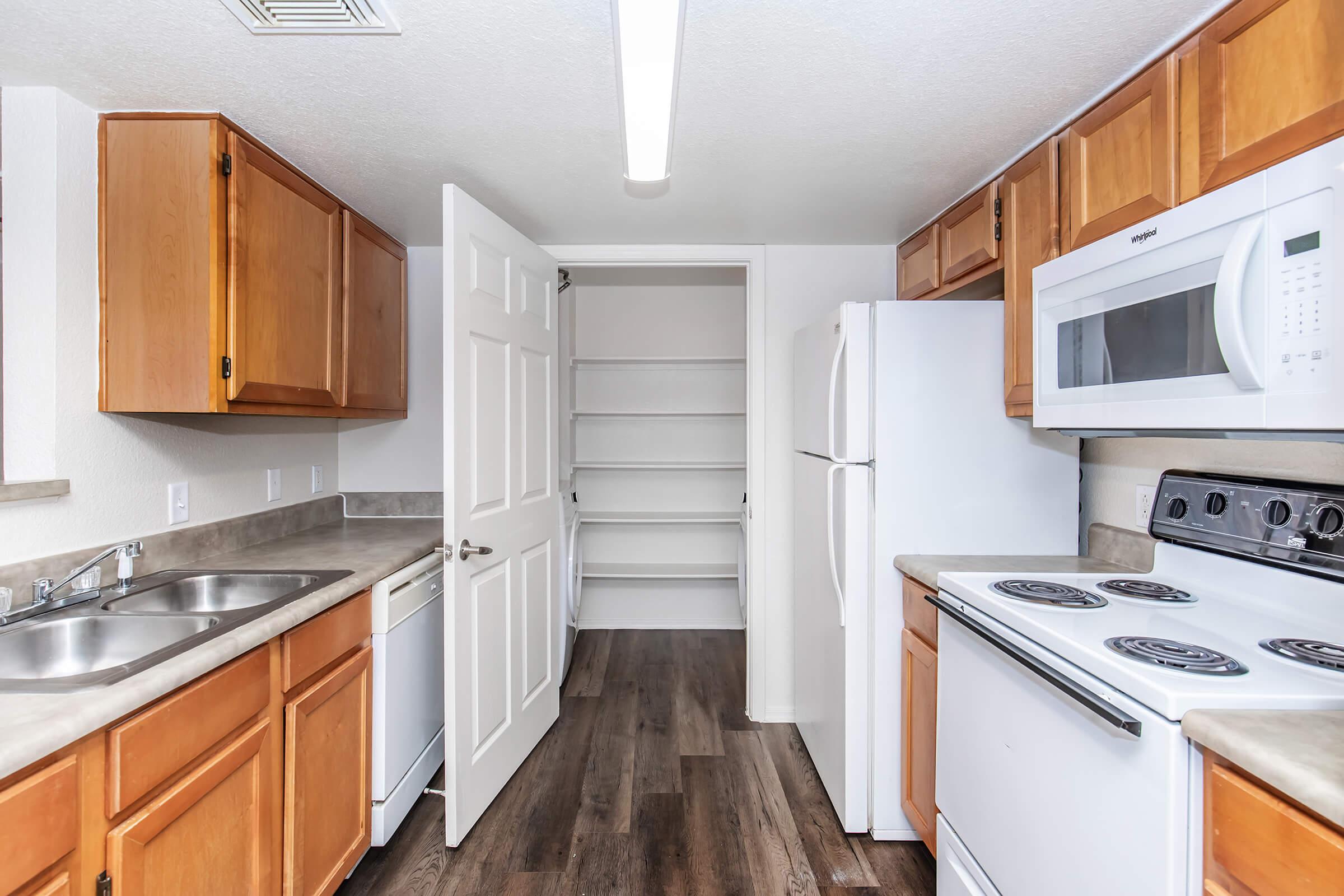 A modern kitchen featuring wooden cabinets, a double sink, and stainless steel appliances. The space includes a white refrigerator, a microwave, and an electric stove. A door leads to a pantry with open shelving, and the floor has dark laminate planks, creating a warm and inviting atmosphere.