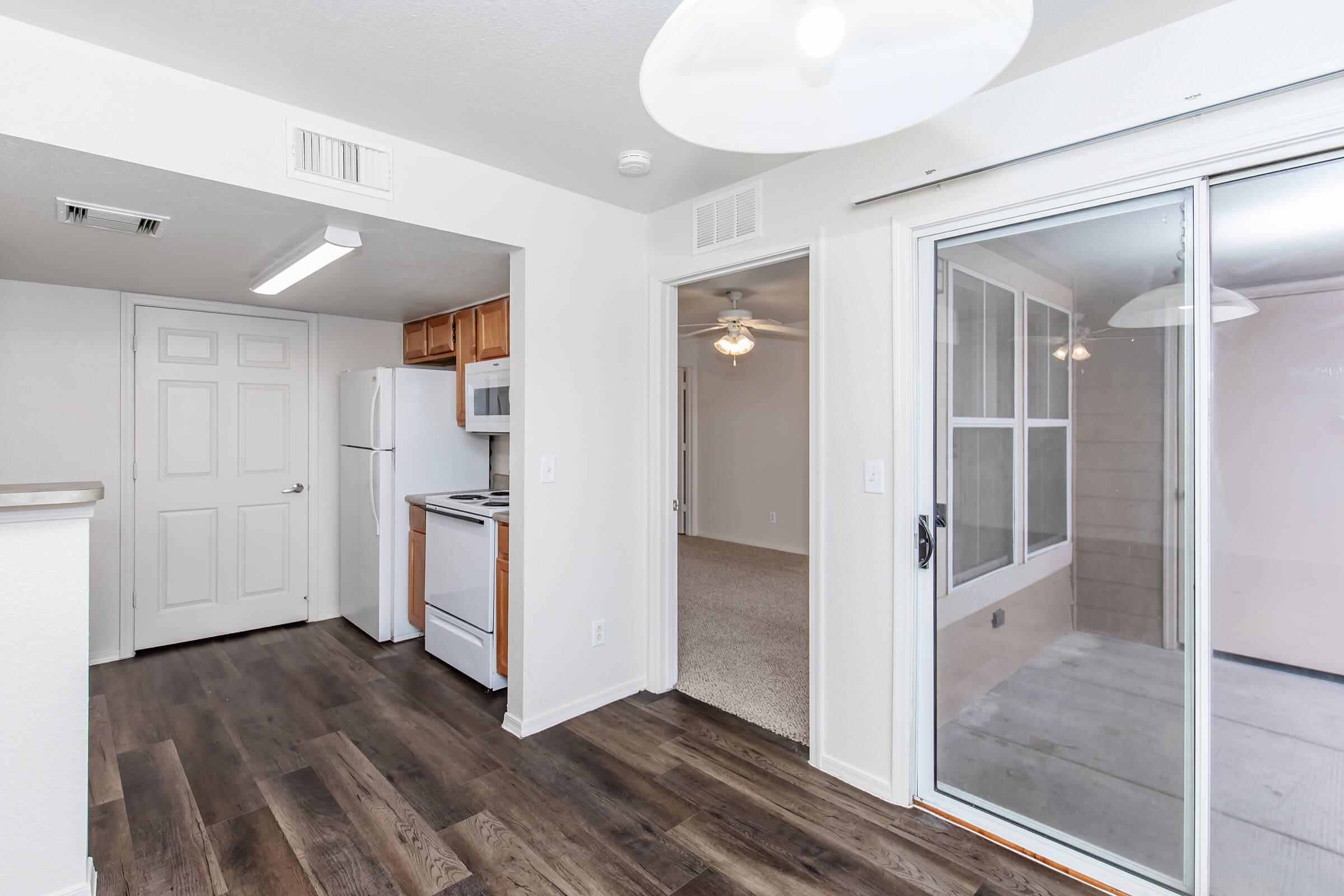 Interior view of an apartment, featuring a kitchen with a refrigerator and stove, an open living space with wood-style flooring, and a sliding glass door leading to a screened patio. The walls are painted light, and there is a ceiling fan in the room.