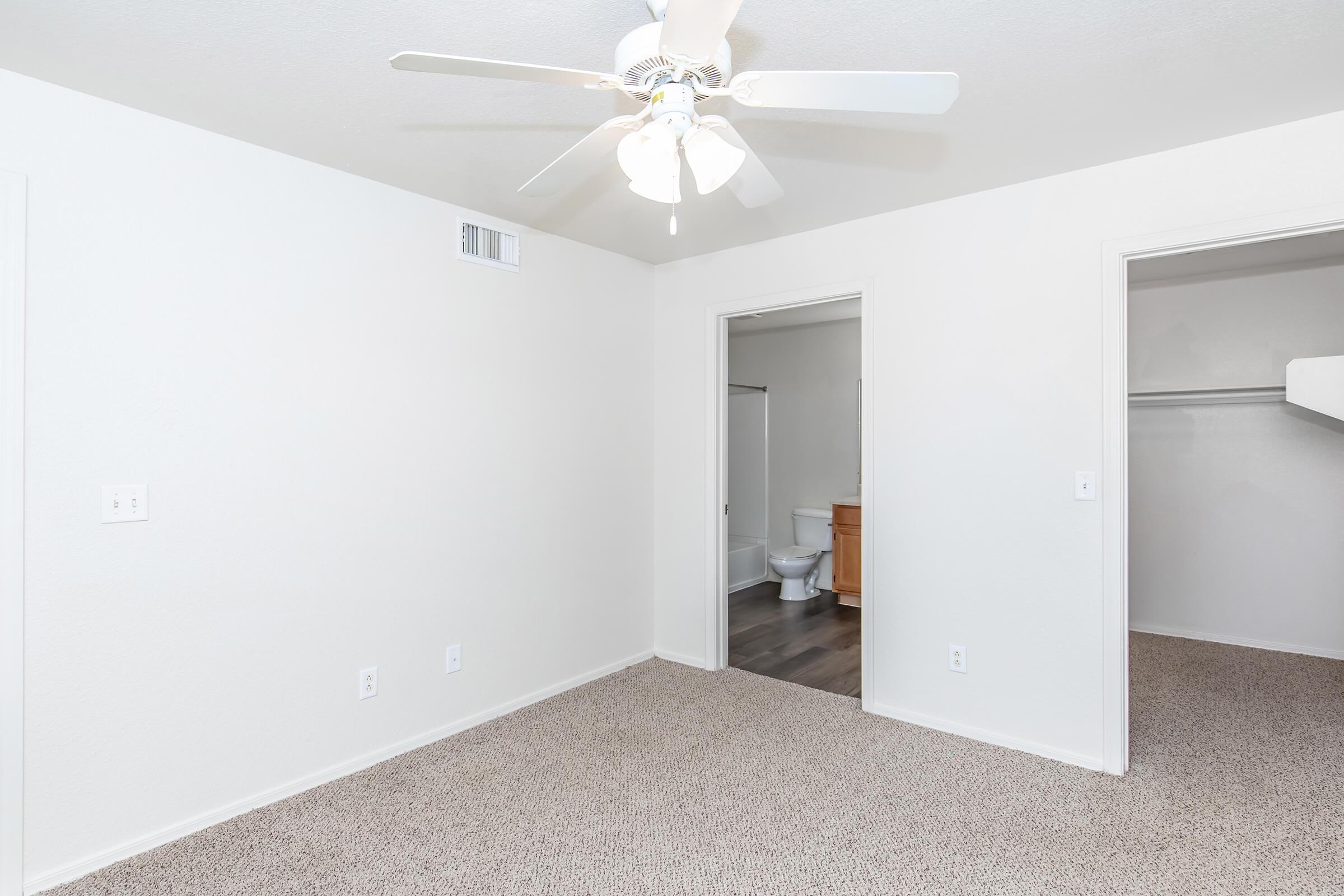 A light and airy room with beige carpet flooring, white walls, and a ceiling fan. There is an open doorway leading to another space, which contains a bathroom and a closet. The overall atmosphere is clean and modern, suitable for various uses.