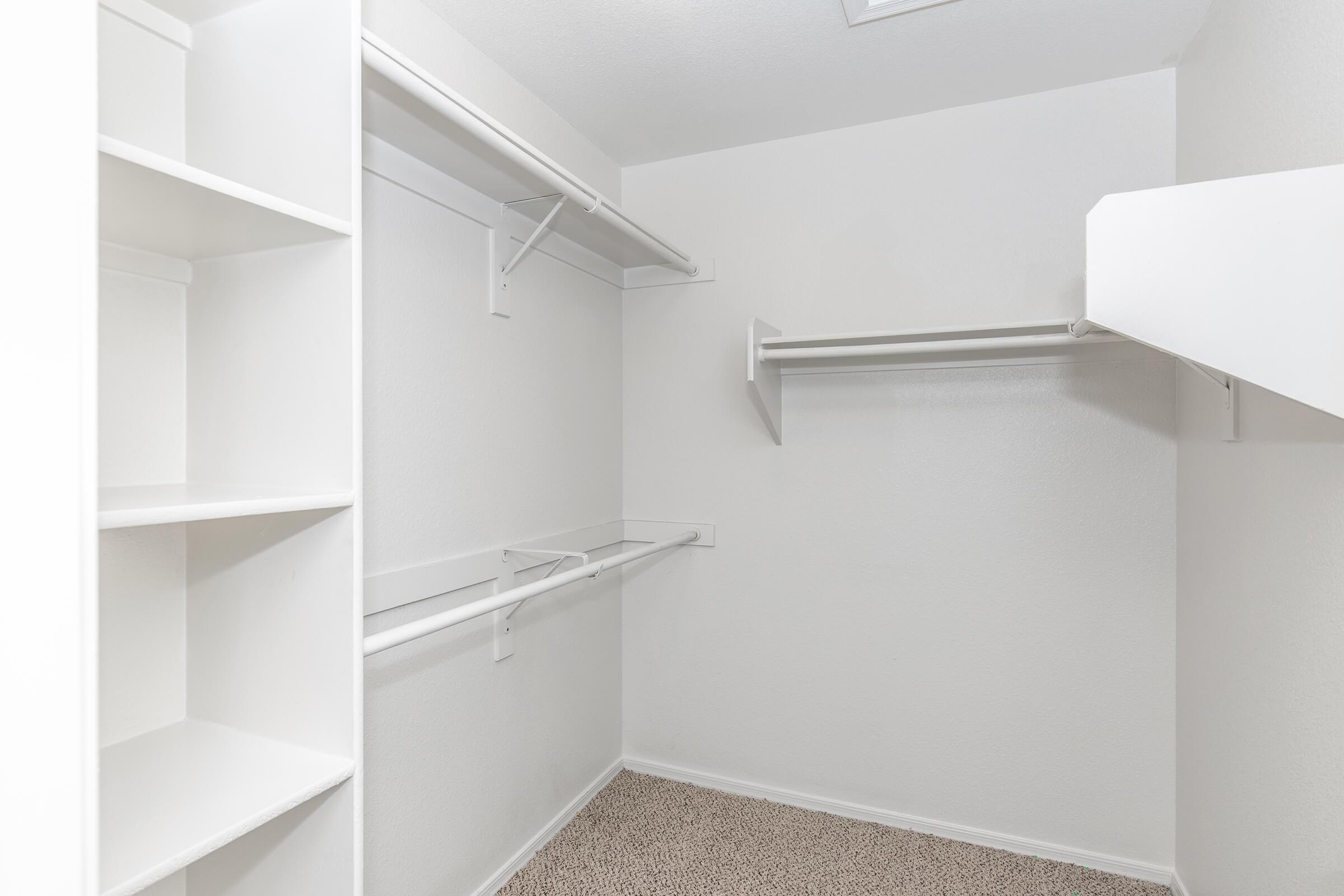A clean, empty walk-in closet featuring white shelving and hanging rods. The walls are painted white, and the floor is carpeted in a neutral shade. The space is well-lit and minimalistic, providing a tidy and organized appearance.