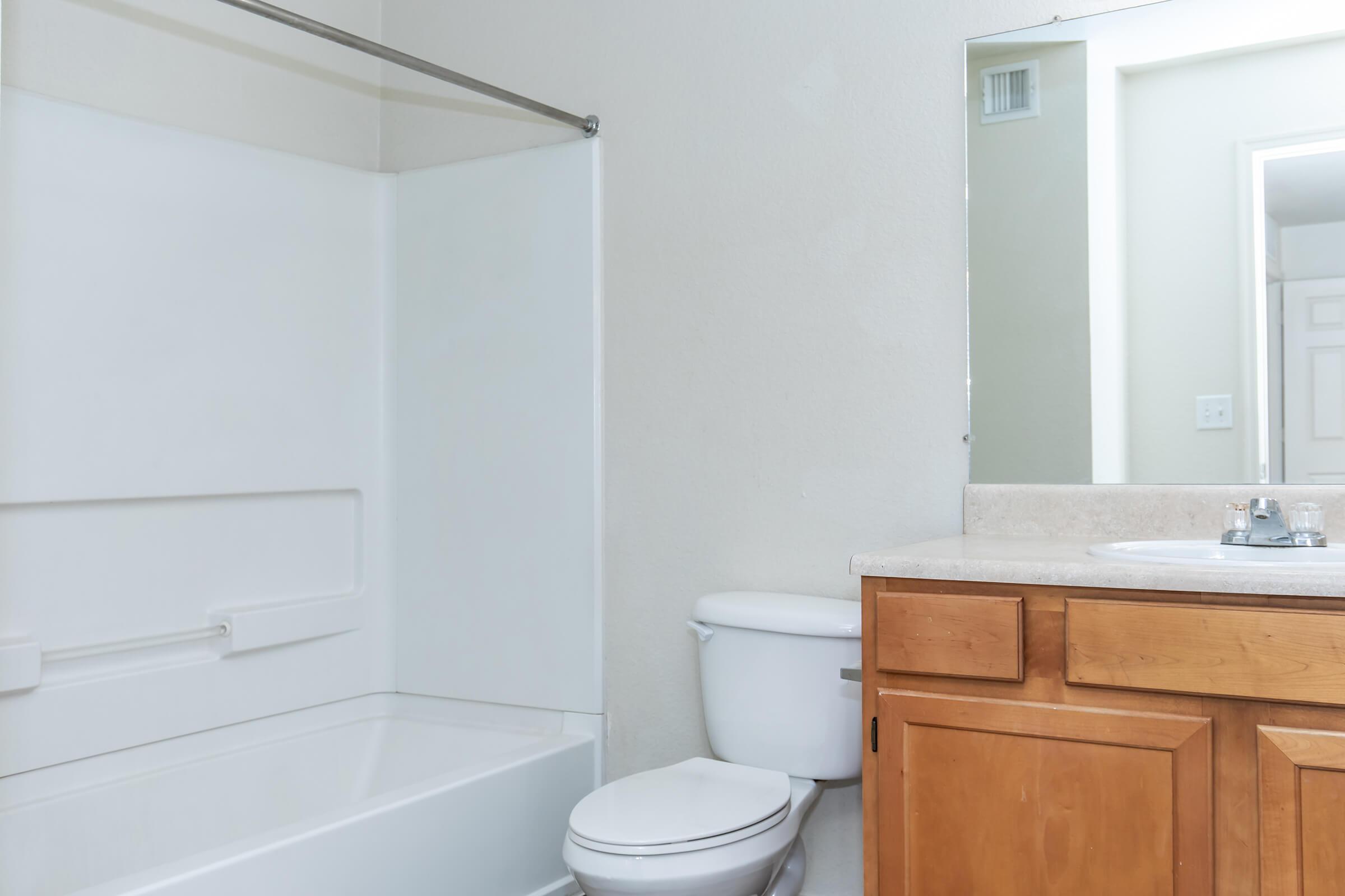 A clean and simple bathroom featuring a white bathtub with a shower curtain, a white toilet, and a wooden vanity with a sink. The walls are light-colored, and there's a large mirror above the sink, providing a bright and spacious feel to the room.