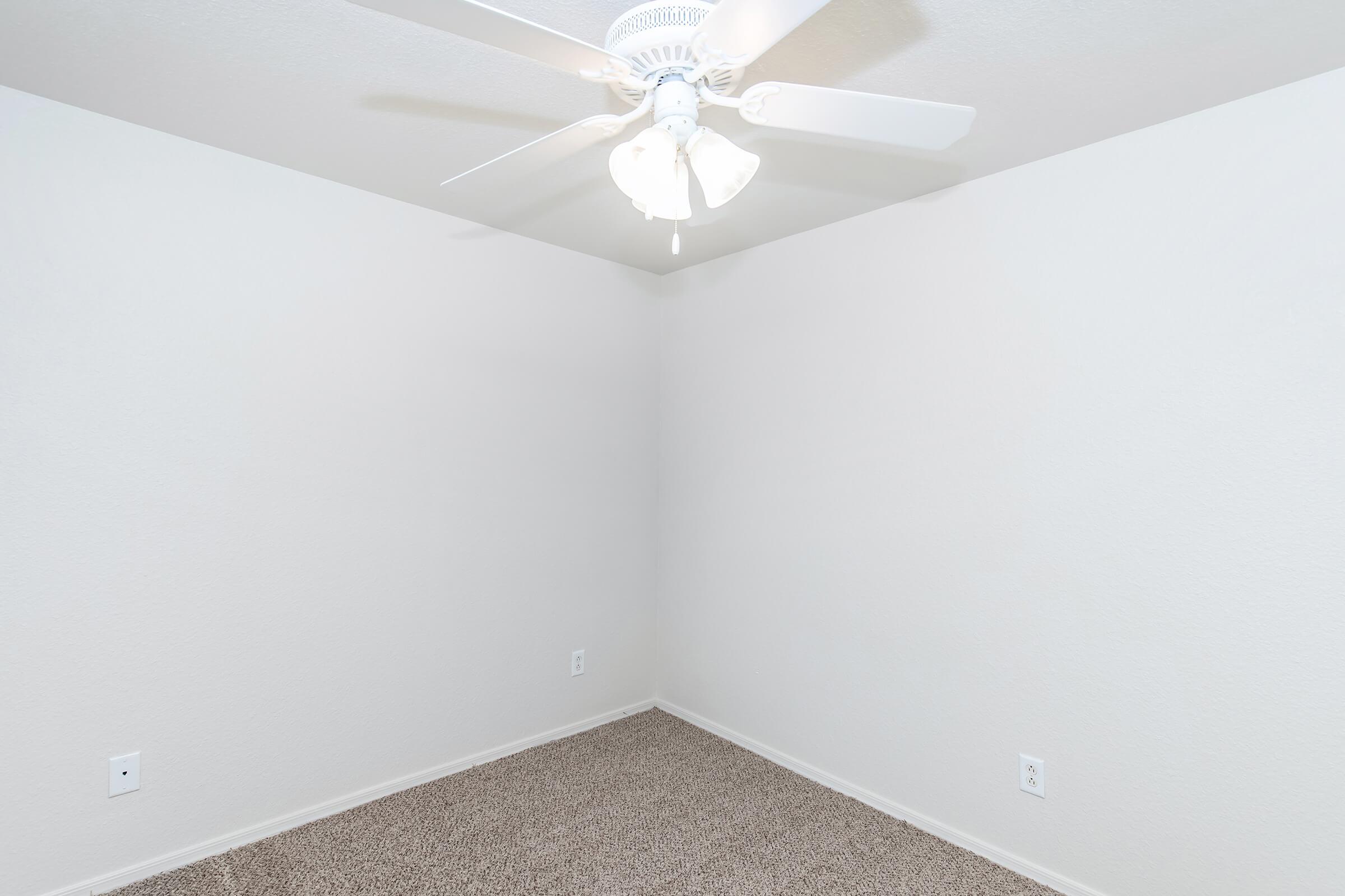 A vacant room with light beige walls and carpeted flooring. The ceiling features a white ceiling fan with light fixtures. There are two electrical outlets visible on the wall, and the overall atmosphere is bright and minimalistic, creating a sense of spaciousness.