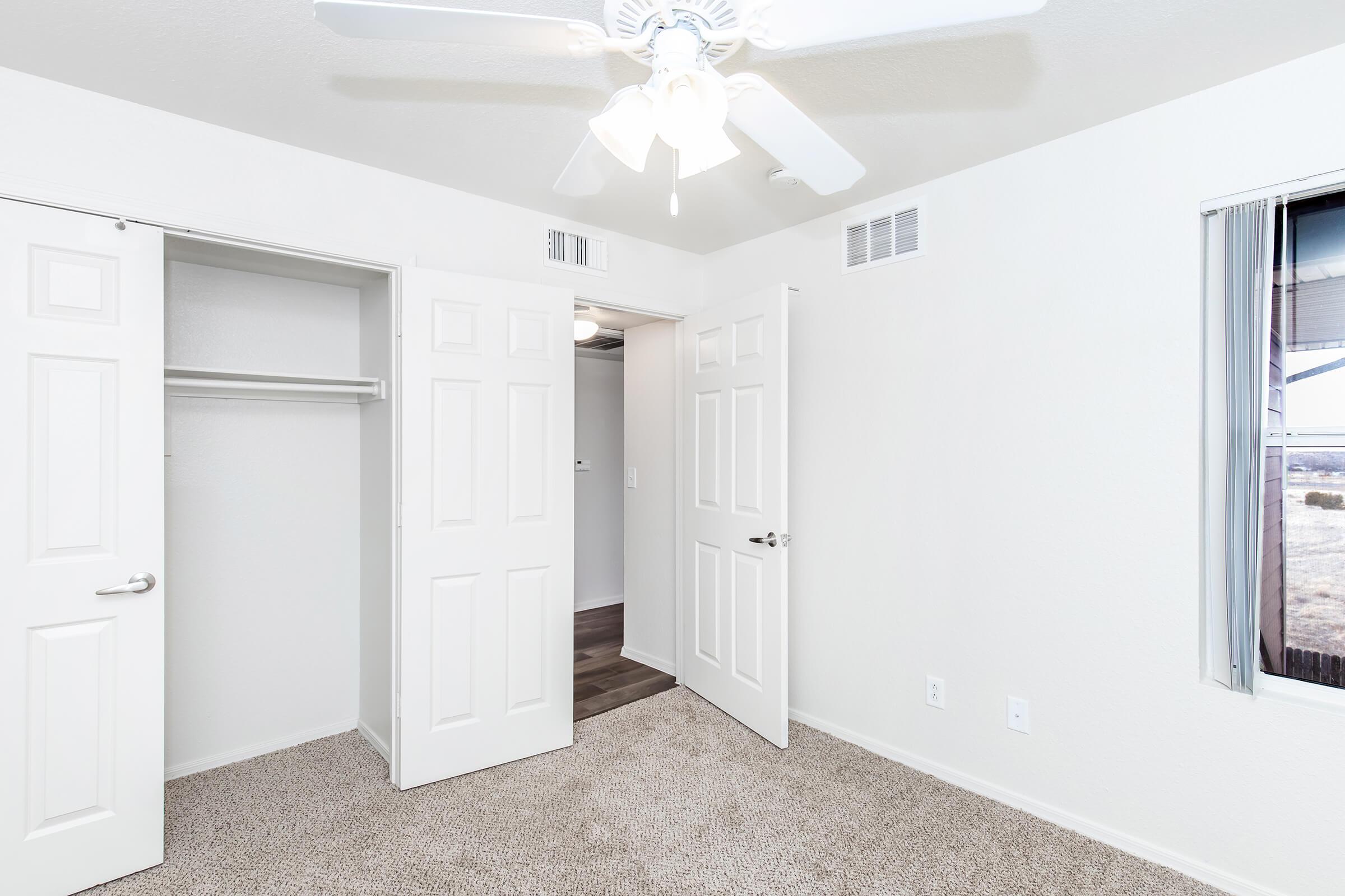 A well-lit, unfurnished room with beige carpet and white walls. There are two open closet doors on the left, and two closed doors on the right. A ceiling fan is installed, and a window is visible, providing natural light. The overall atmosphere is clean and minimalist.
