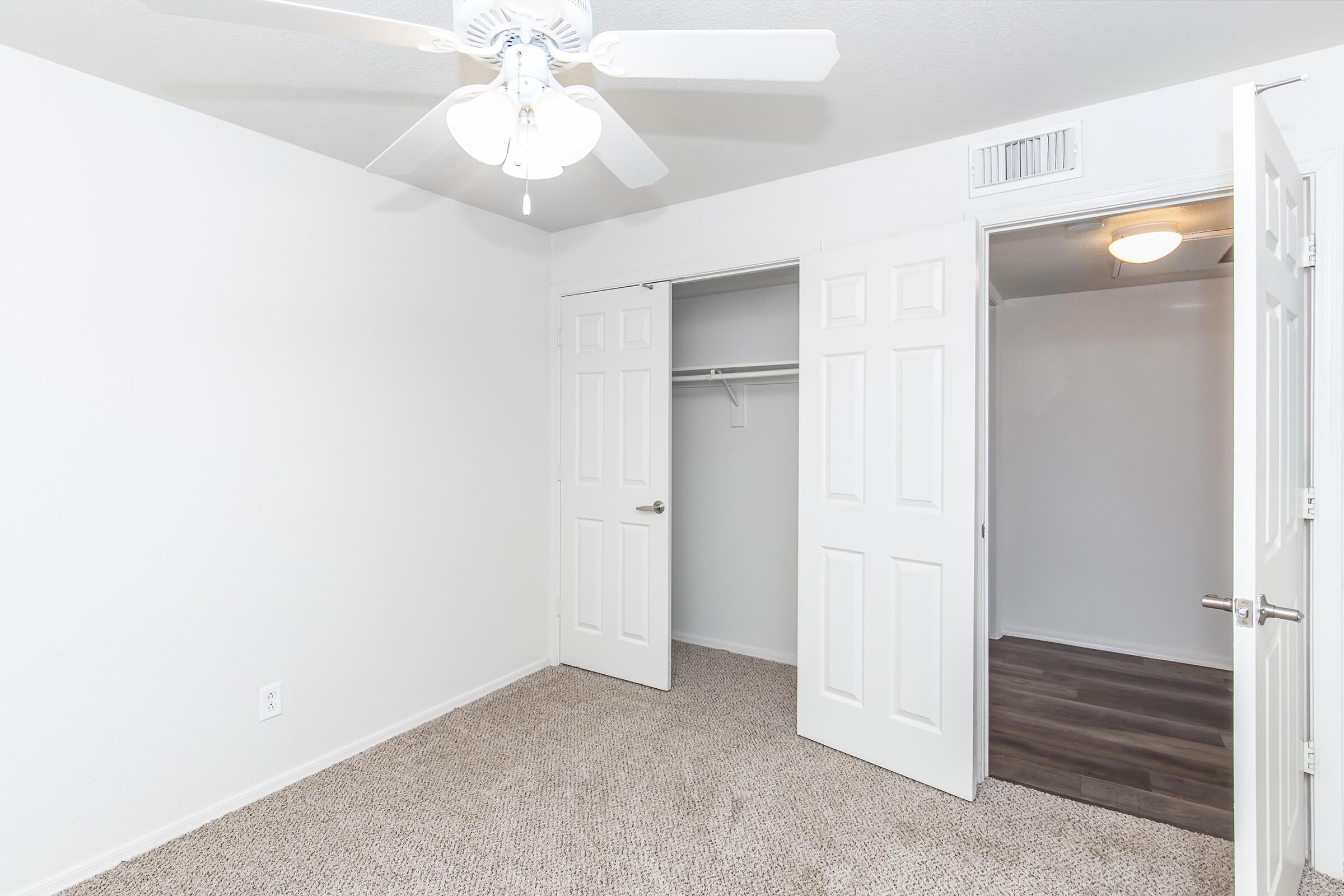 A spacious, well-lit bedroom featuring neutral-colored walls and plush carpeting. There are two white double closets with sliding doors, a ceiling fan with light fixture, and a door leading to another room. The overall atmosphere is clean and inviting, perfect for personalizing.