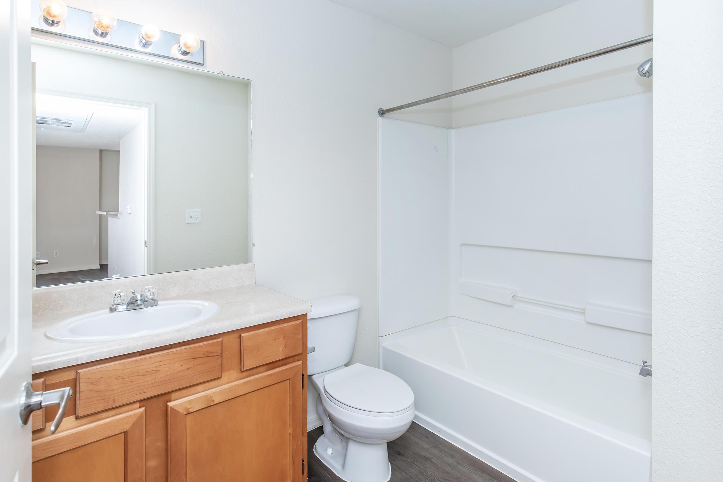 A clean and simple bathroom featuring a beige countertop with a sink, a white toilet, and a bathtub with a shower. A large mirror above the sink reflects the light from overhead fixtures. The walls are painted a light color, enhancing the spacious feel of the room.