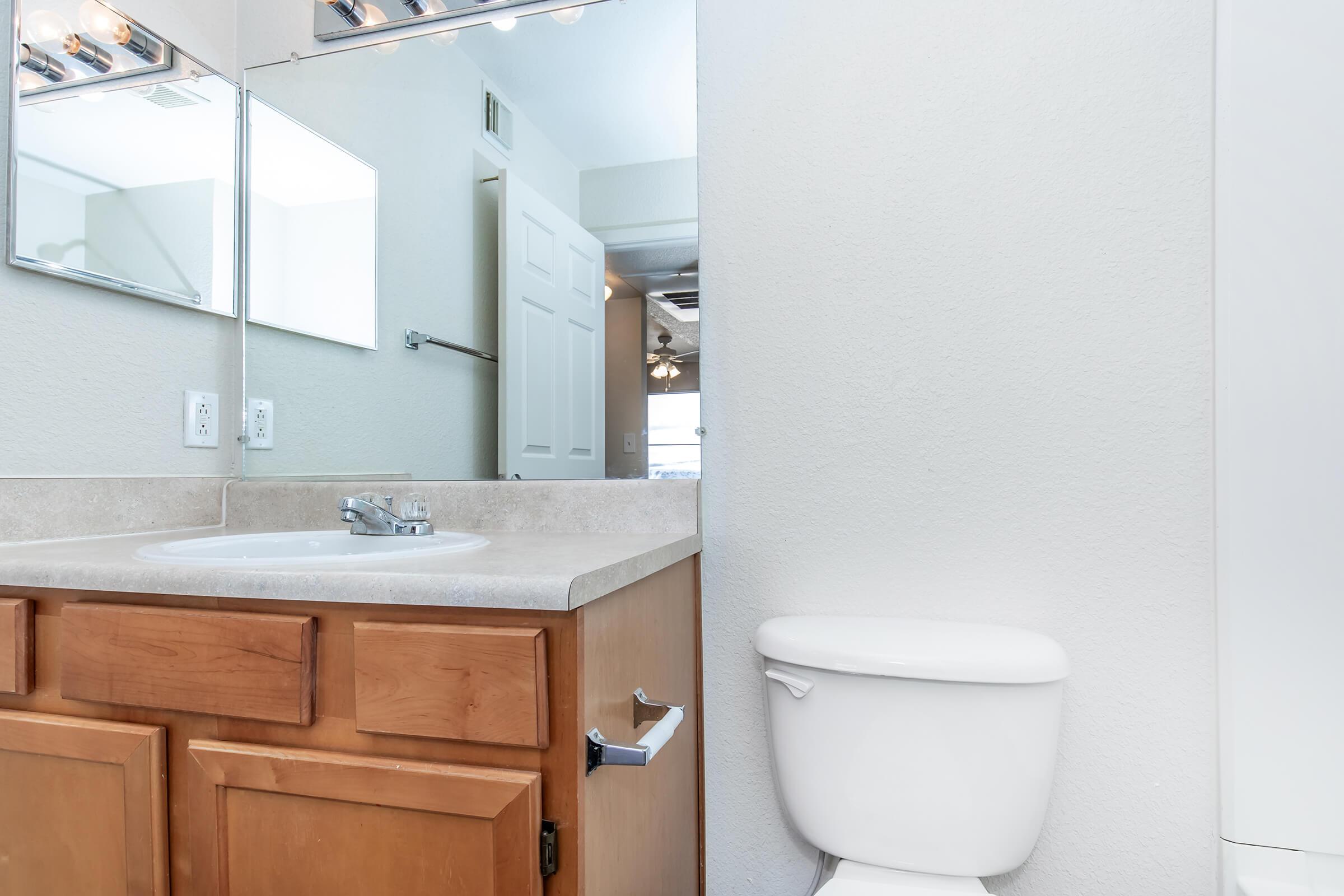 A clean, bright bathroom featuring a light-colored countertop with a sink, wooden cabinetry, and a white toilet. Mirrors reflect the space, and a door leads to another area. The walls are painted in a light tone, enhancing the brightness of the room.