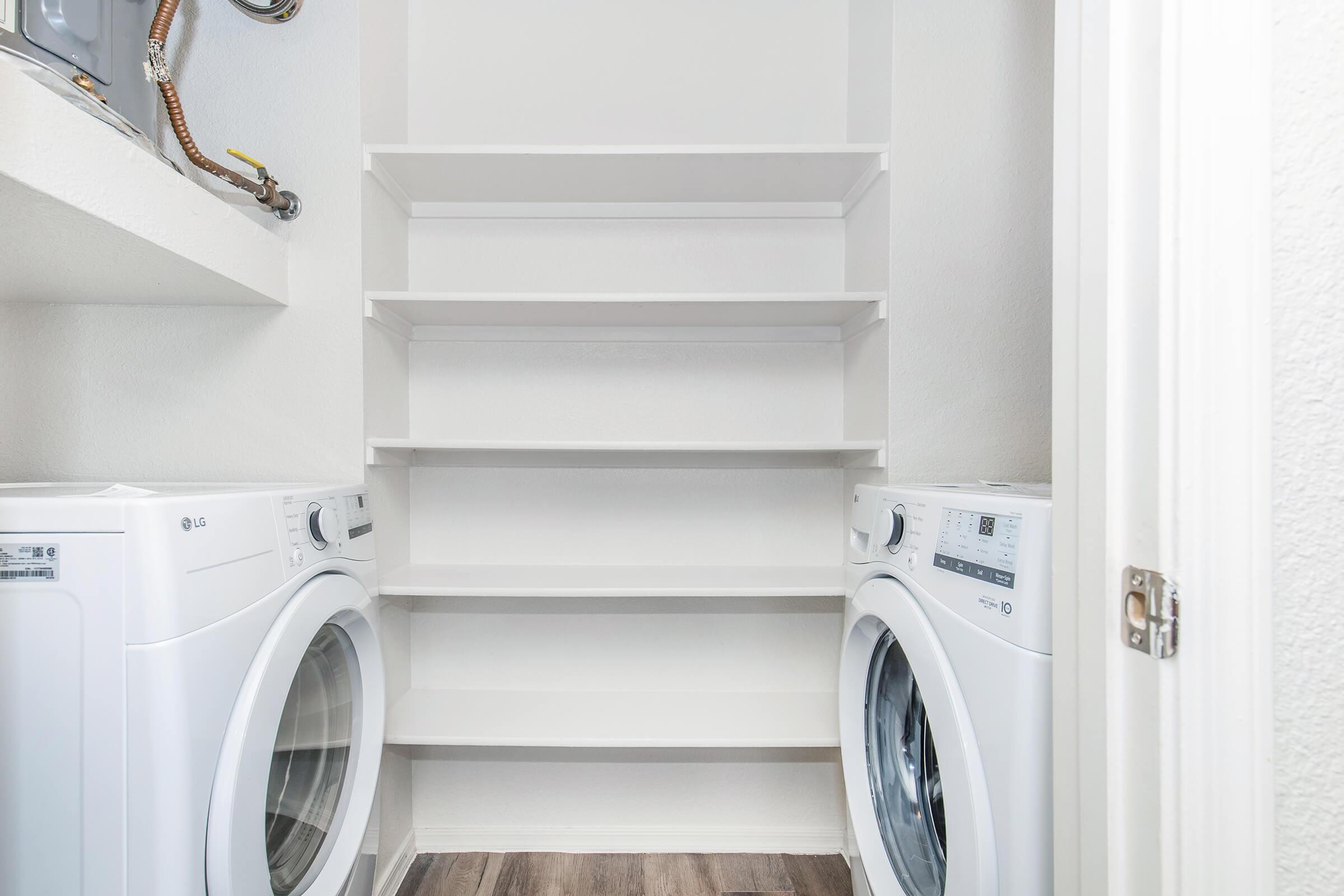 A clean laundry room featuring two white washing machines side by side. Above them is a shelf for storage, and the walls are painted in light colors. The flooring is made of wood, and there are utility connections visible on the left side of the image.