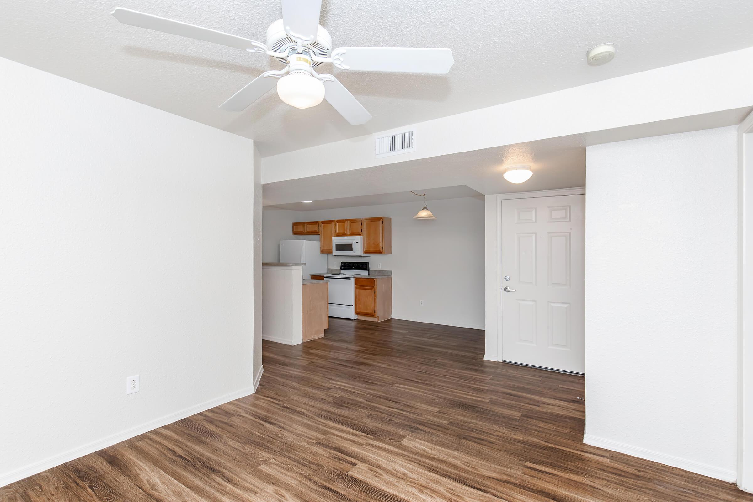 Interior view of a modern apartment featuring a living area with wooden flooring, a ceiling fan, and bright walls. The open layout connects to a kitchen area with white appliances, light wood cabinets, and a small dining space. The entrance door is visible along with an overhead light fixture.