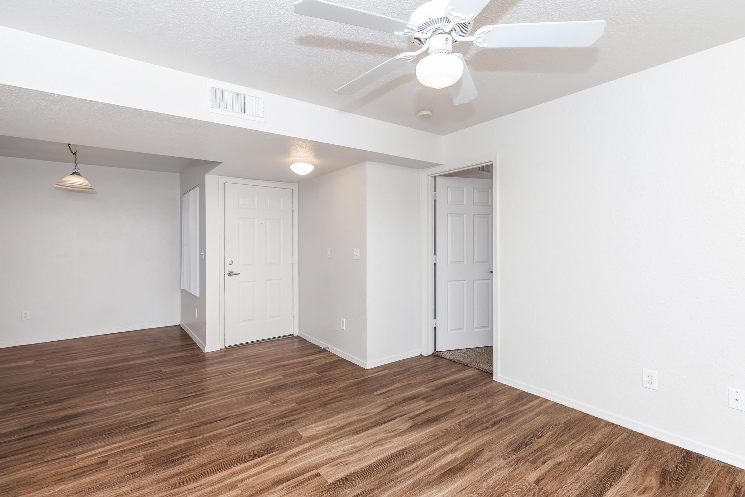 A spacious, well-lit room featuring light-colored walls and wood laminate flooring. Two doorways lead to adjacent rooms, while a ceiling fan hangs above. A light fixture is visible in the corner, adding to the minimalistic decor. The overall atmosphere is clean and inviting.