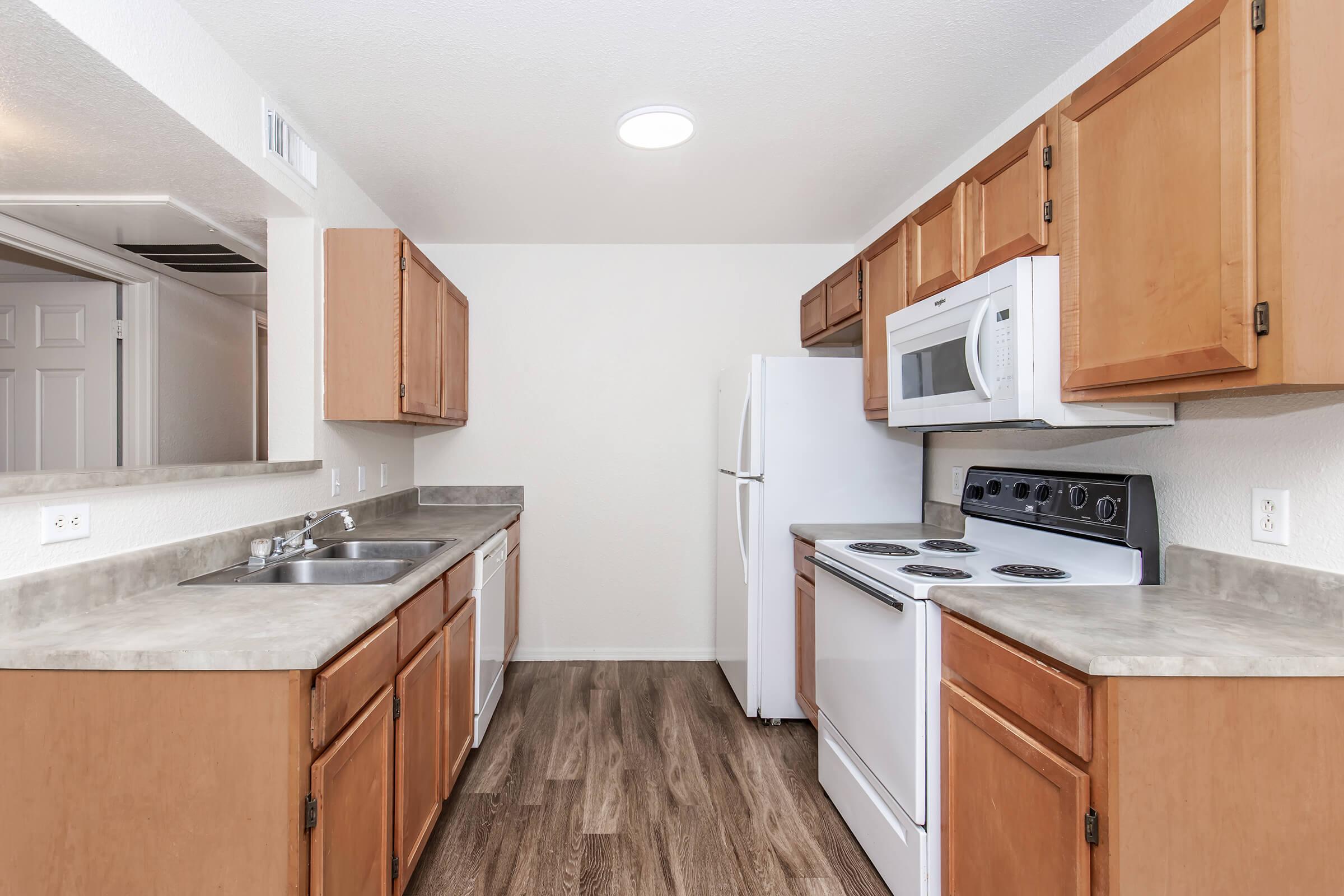 Modern kitchen featuring light wood cabinets, a gray countertop, and stainless steel appliances. The layout includes a double sink, a microwave, an electric stove, and a refrigerator. The space is well-lit, highlighting the open and clean design with laminate flooring.