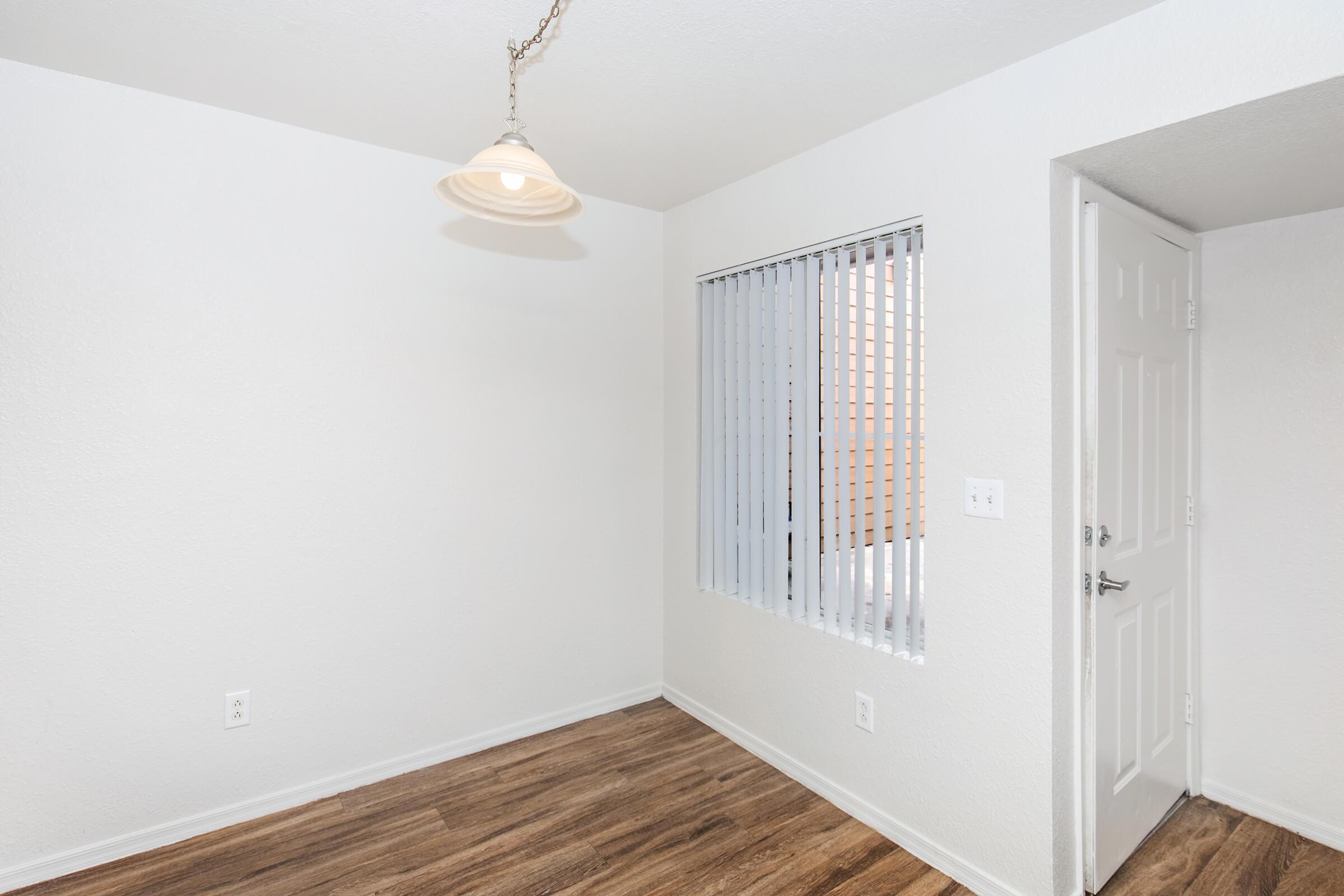 A well-lit room with white walls and wooden flooring. There is a ceiling light fixture, and a window with vertical blinds allowing natural light to enter. A doorway is visible on the right side, leading to another space. The overall atmosphere is clean and minimalistic.
