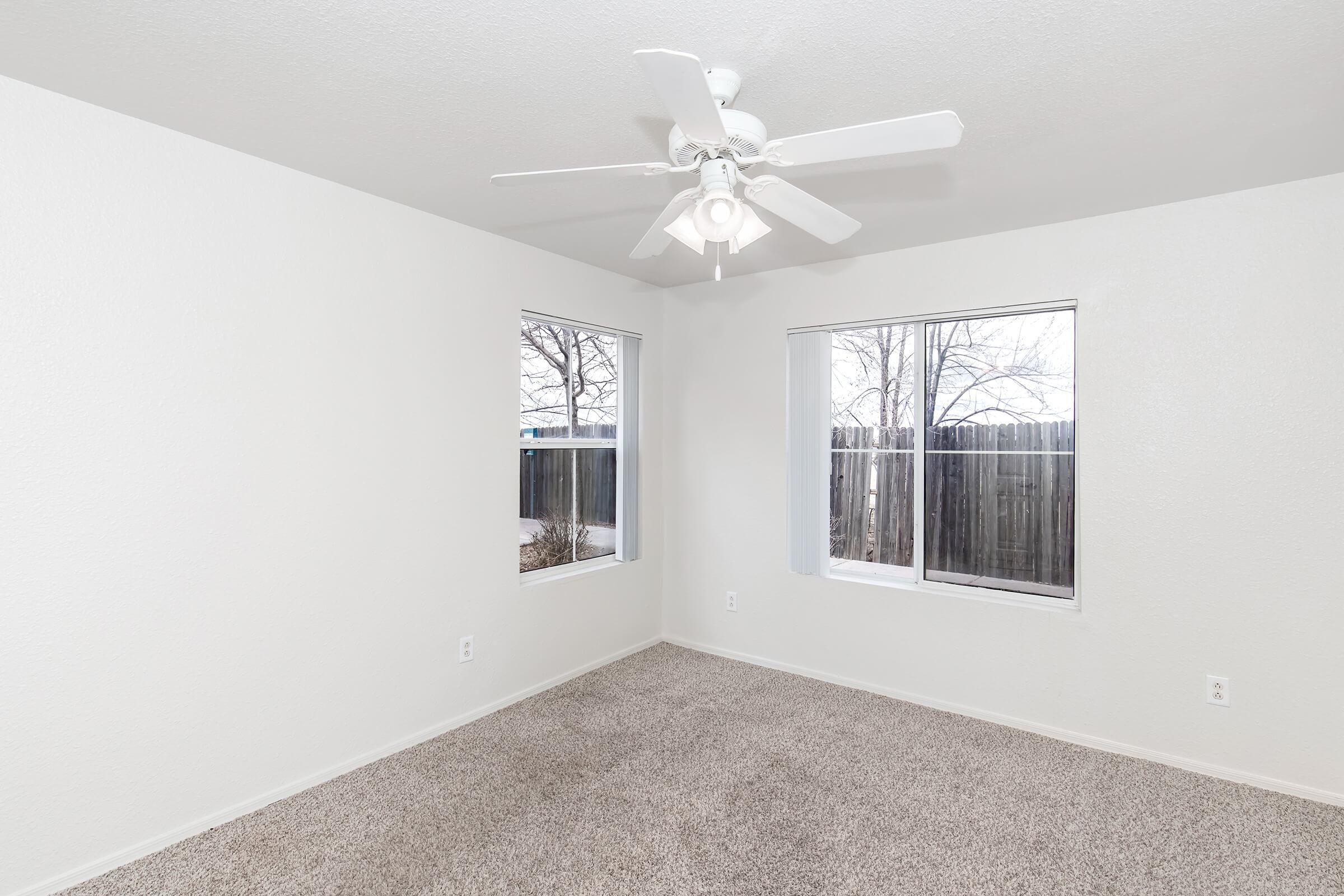 A well-lit empty room with light-colored walls and soft carpet. It features two windows providing natural light, and a ceiling fan mounted in the center. The atmosphere is airy and spacious, suitable for various interior design possibilities. A partially visible backyard adds a touch of greenery.