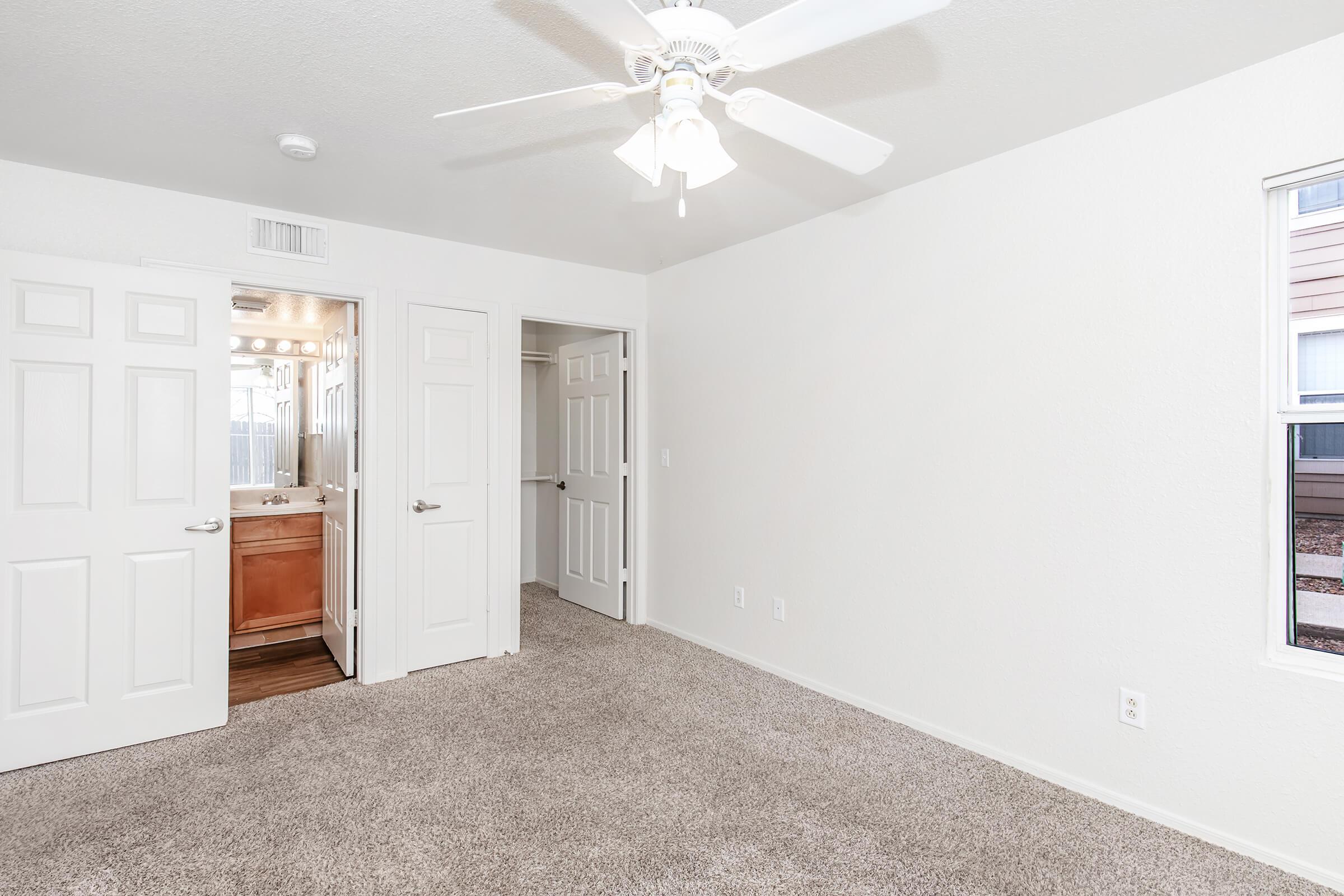 A spacious room with light-colored walls and plush carpeting. A ceiling fan is mounted above. There are two open doors: one leading to a bathroom with a vanity, and the other to a hallway. A window allows natural light to enter, illuminating the clean, uncluttered space.