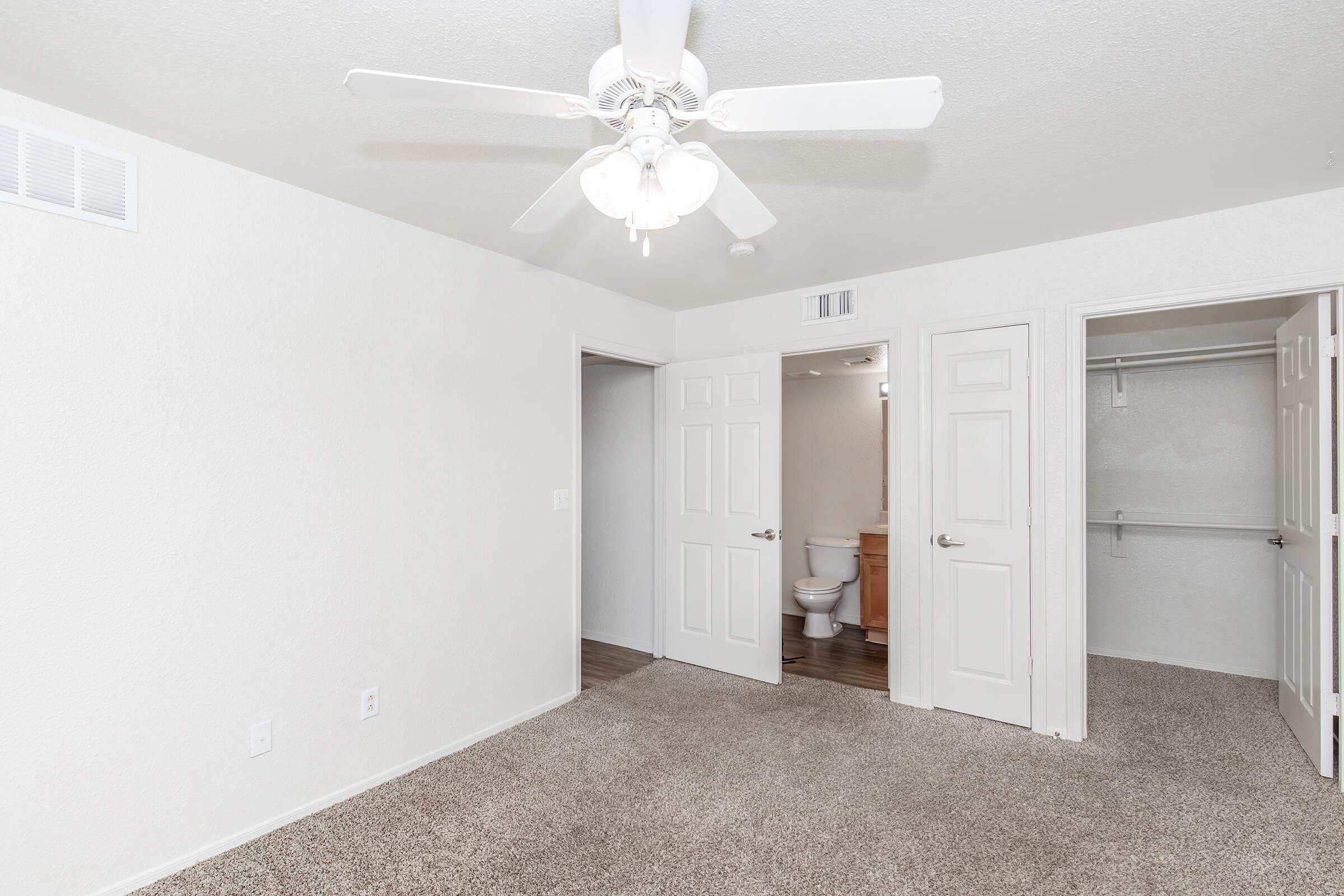 A well-lit, neutral-colored bedroom featuring a ceiling fan, carpeted flooring, and two doors leading to a bathroom and a closet. The room is spacious and has a light, airy feel, ideal for comfortable living.