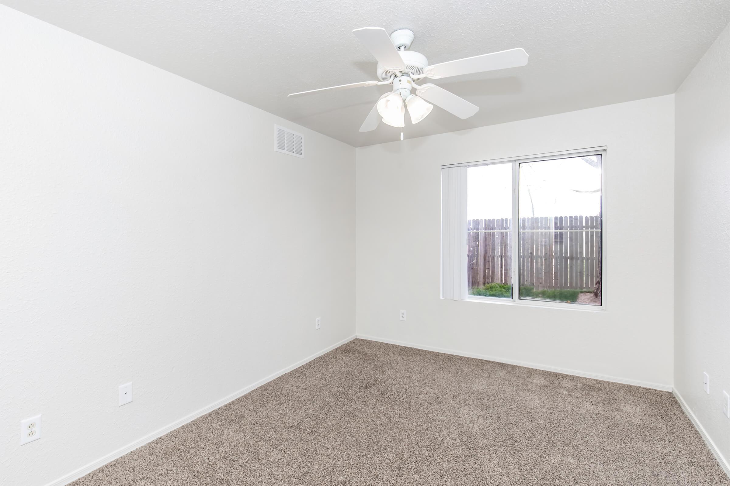 Empty room with light-colored walls and a ceiling fan. The floor is covered in beige carpet, and there is a window with white blinds, overlooking a fenced outdoor area. The room appears spacious and well-lit.