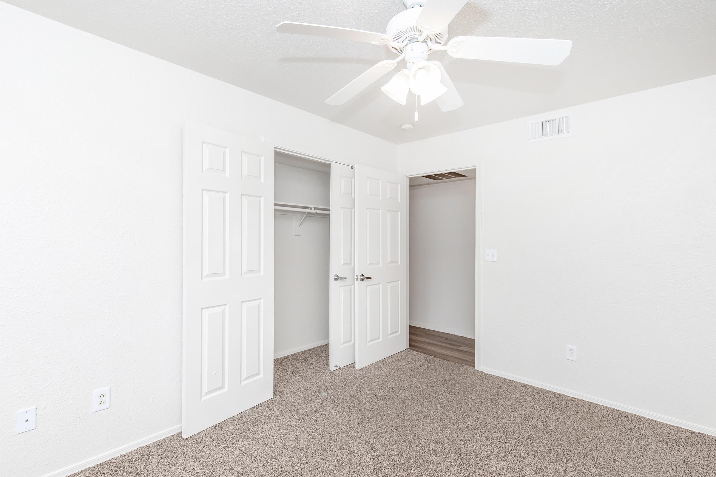 A spacious room with beige carpet, featuring two open closet doors, a ceiling fan with light, and light-colored walls. The room has a small doorway leading to another space, creating a bright and airy atmosphere. Ideal for use as a bedroom or office.