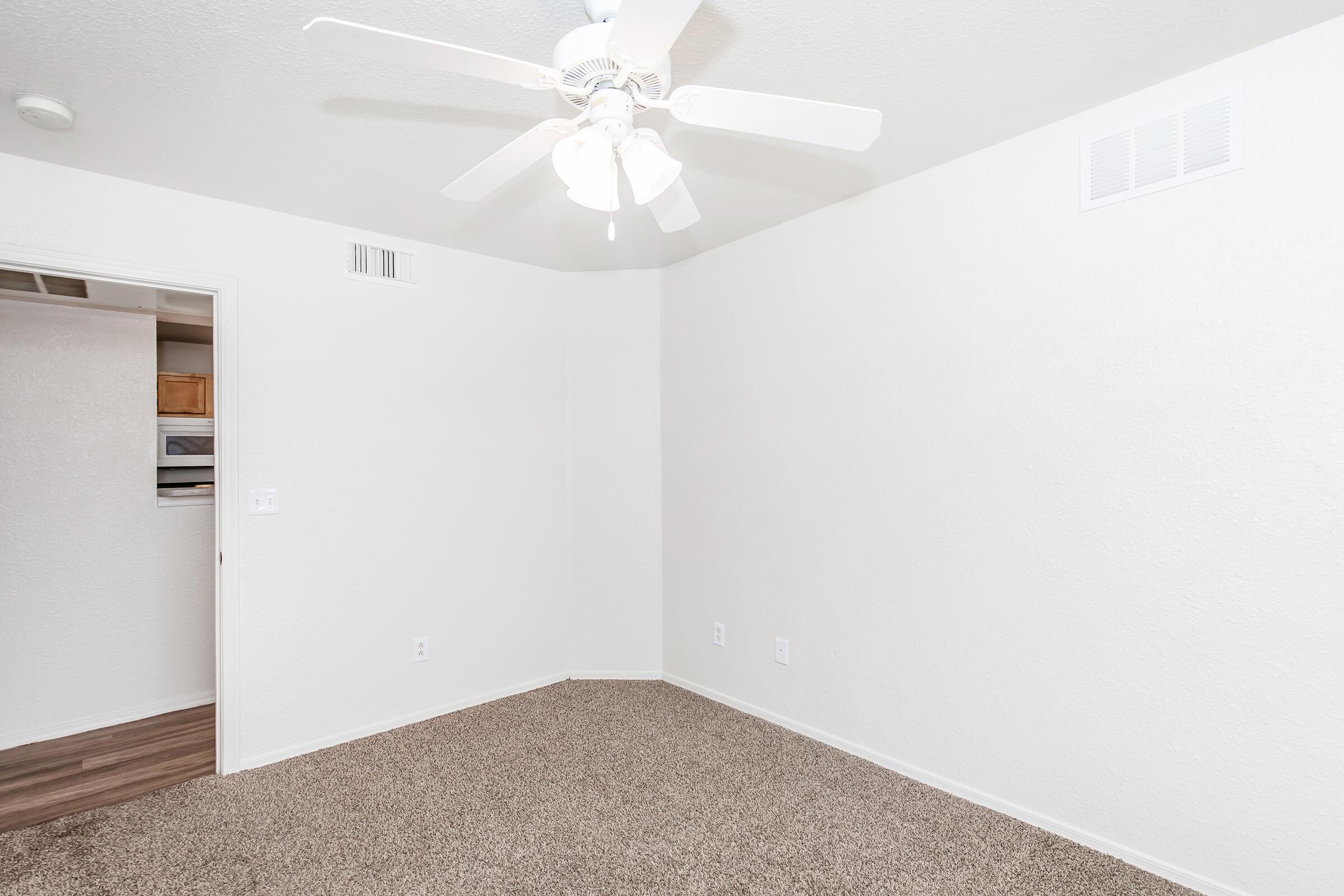 A clean, empty room featuring light-colored walls and a ceiling fan. The floor is covered in carpet, and there is a closet visible in the background. Natural light is suggested by the bright, neutral color scheme, creating a spacious and inviting atmosphere.