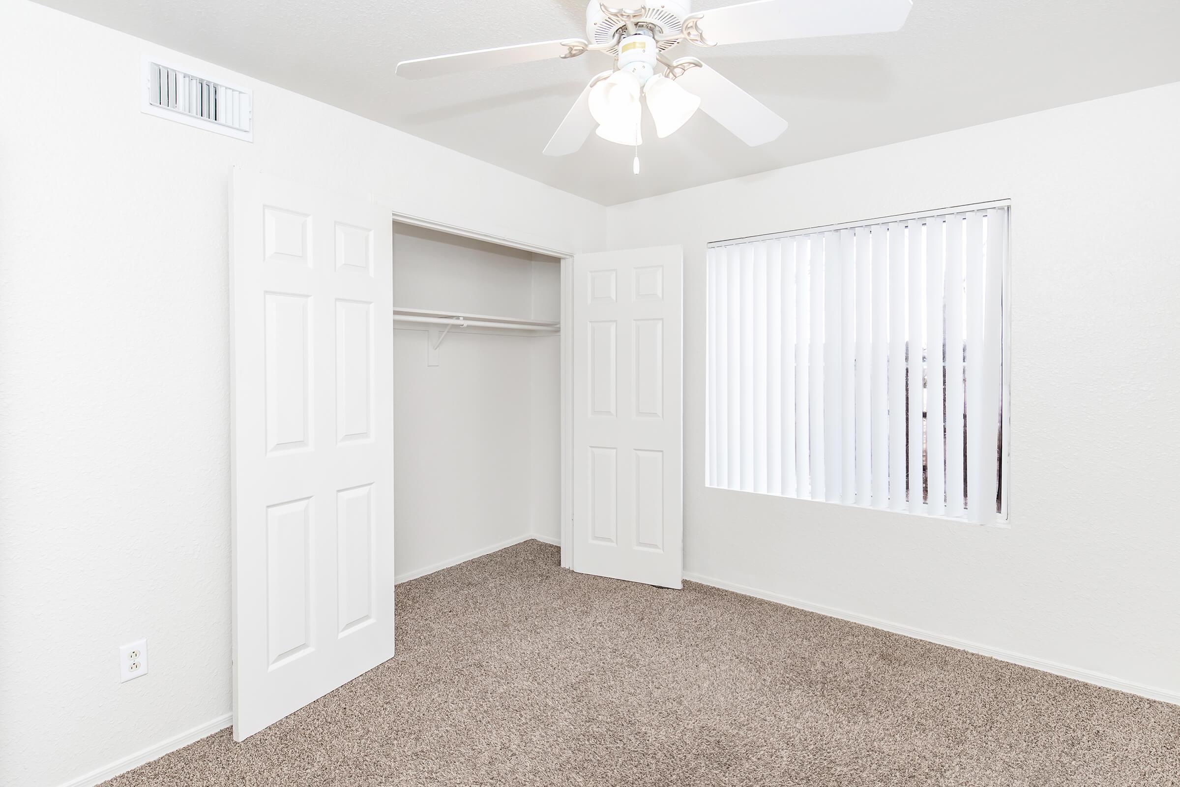 A bright, empty room with light beige carpet and white walls. It features a ceiling fan, a window with vertical blinds, and an open closet with double doors. The room has a clean and spacious feel, suitable for various furnishings.