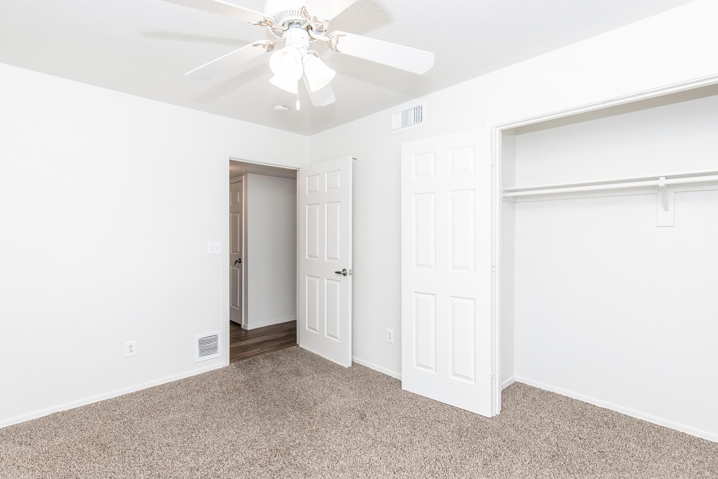 A well-lit empty room with beige carpet, featuring a ceiling fan and white walls. There's an open closet to the right and two doors: one leading to another room and the other possibly a bathroom or closet. The space appears clean and modern, ideal for various uses.