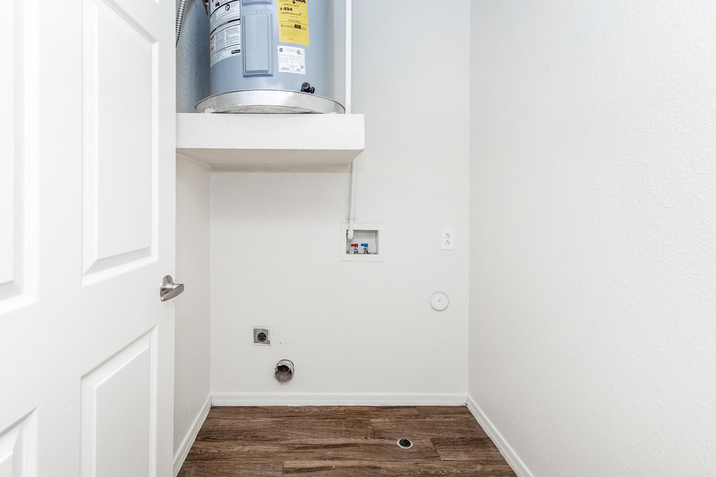 A small utility room featuring a water heater mounted on a shelf above the floor. The walls are painted white, and the floor is covered with wooden laminate. There are visible plumbing connections on the wall and a drain in the floor, with a closed door leading to the room.