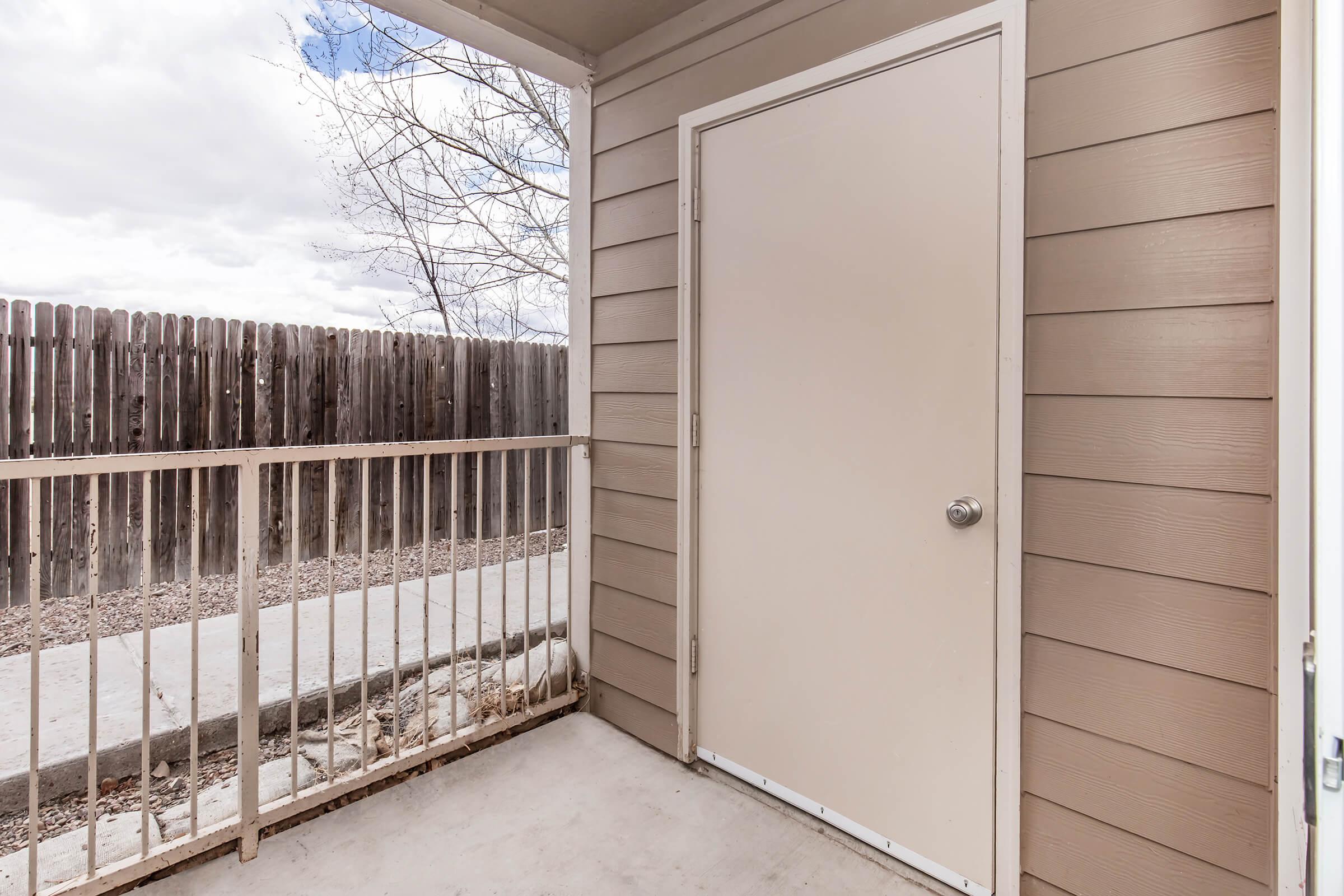 A porch area featuring a beige door on the left side, wood paneling on the walls, and a metal railing. In the background, there is a wooden fence and a cloudy sky. The concrete floor is visible, leading to a path. The overall setting appears to be a residential outdoor space.