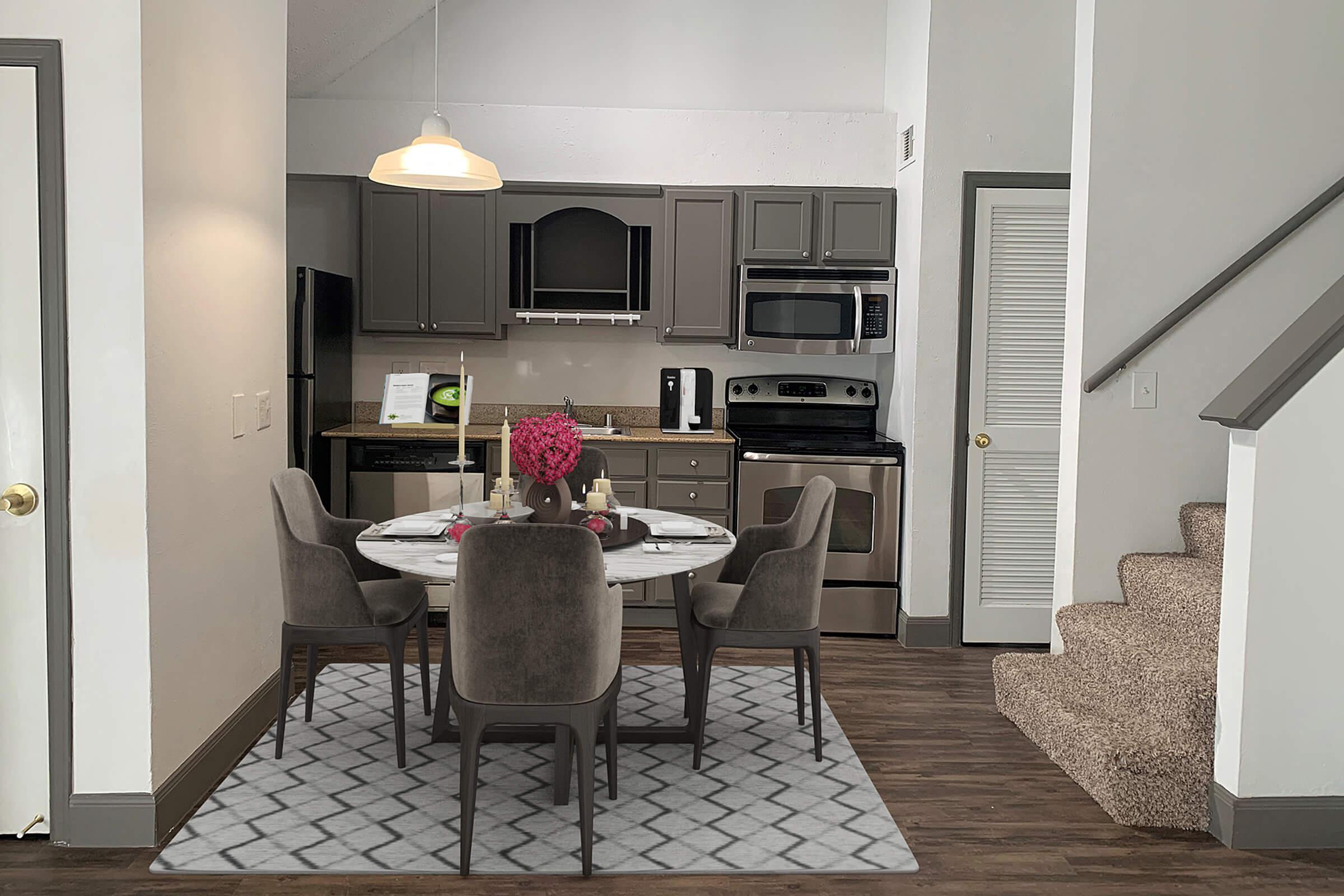 A modern kitchen and dining area featuring a round marble table with four gray chairs, a centerpiece of pink flowers, and a gray patterned rug. The kitchen has dark gray cabinets, stainless steel appliances, and a staircase leading to an upper level. Natural light is coming from a ceiling light fixture.