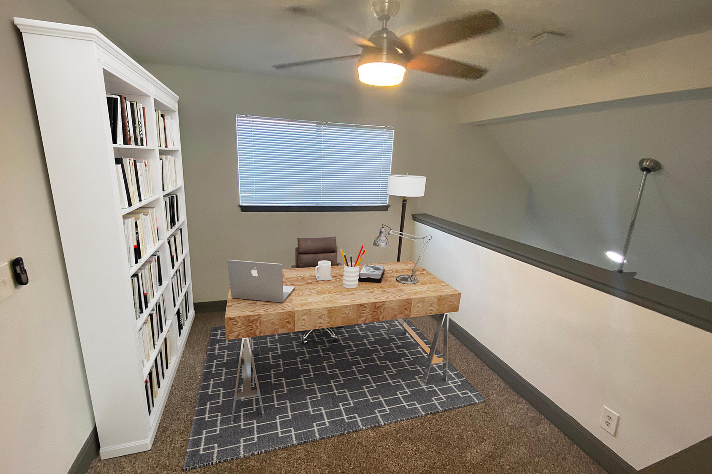 A modern home office featuring a wooden desk with an open laptop, a white lamp, and stationery. Behind the desk is a comfortable brown chair. A white bookshelf filled with books stands against one wall, while a textured gray rug covers the floor. A ceiling fan and a window with blinds provide a bright and airy atmosphere.