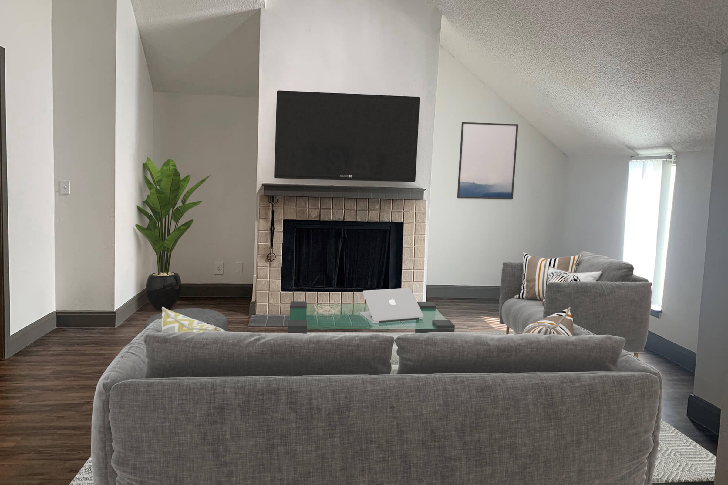 A modern living room with a high, sloped ceiling and a flat-screen TV mounted above a stylish fireplace. Two gray sofas face each other, with a green coffee table in the center. A potted plant stands in the corner, and natural light streams through two windows with light curtains. A minimalistic art piece hangs on the wall.