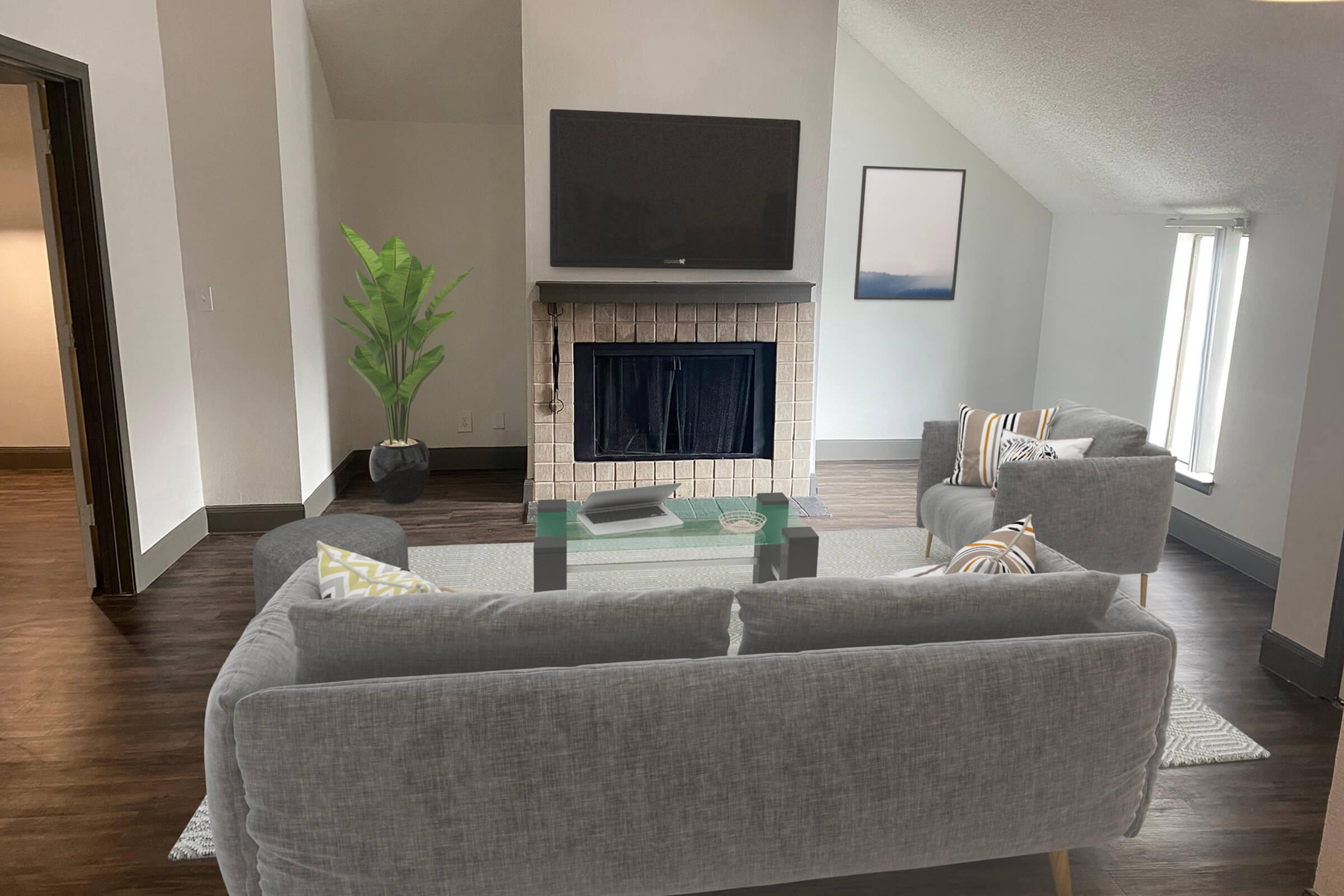 A cozy living room featuring a grey fabric sofa and two matching chairs, a glass coffee table in the center, and a fireplace with a TV mounted above it. Natural light streams in through two tall windows, and a potted plant adds a touch of green to the minimalist decor. Modern and inviting ambiance.