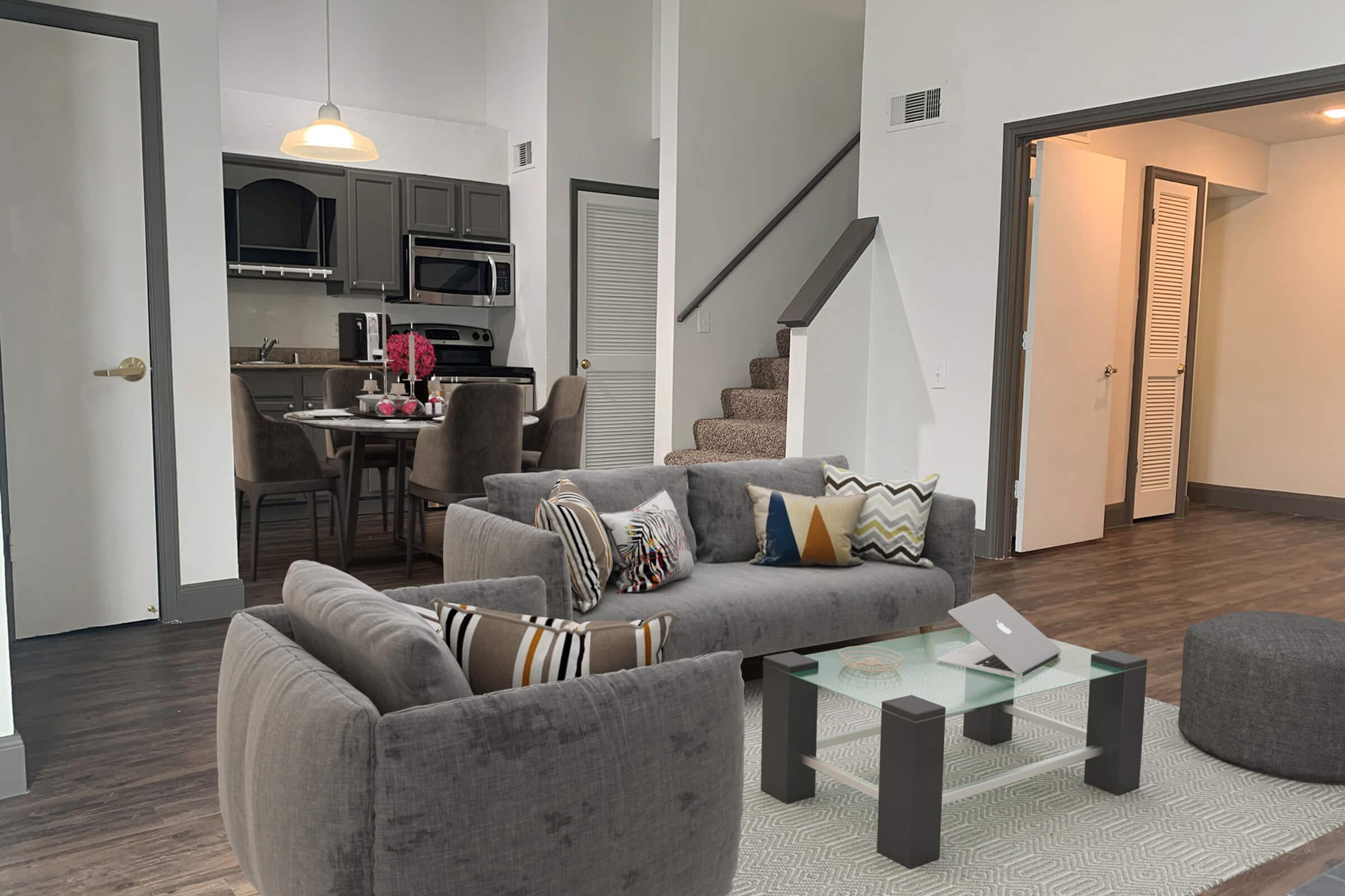 A modern, open-concept living space featuring a gray sofa with patterned cushions, a glass coffee table, and a cozy area rug. In the background, a kitchen with dark cabinets and appliances is visible. A dining table with chairs is set beside the kitchen, and a staircase leads to an upper level. Natural light fills the room.