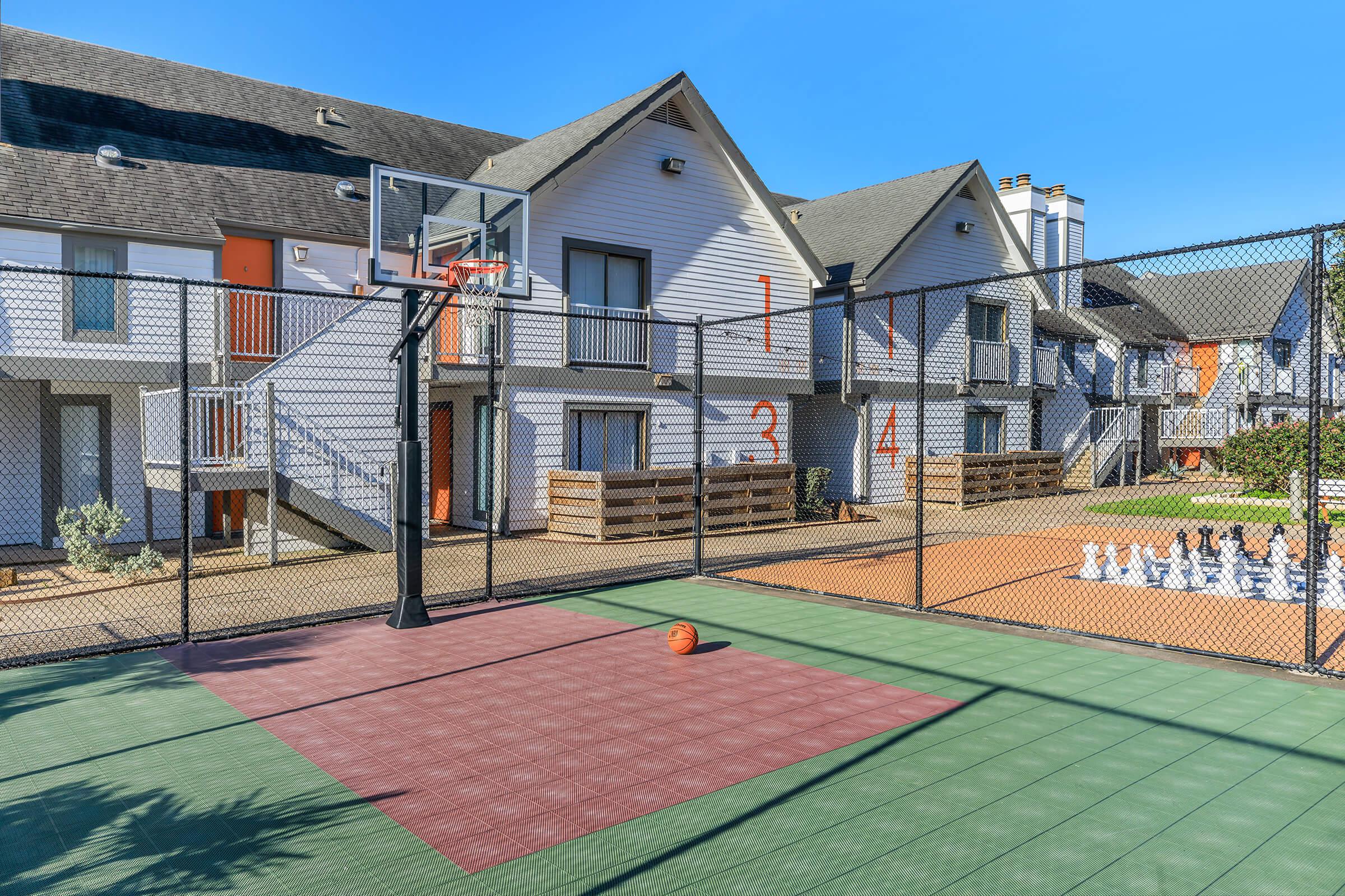 A basketball court with a hoop and a bright orange basketball in the foreground. In the background, there are several apartment buildings with numbered doors, surrounded by greenery. The scene is sunny and features a fenced area, creating an inviting recreational space.