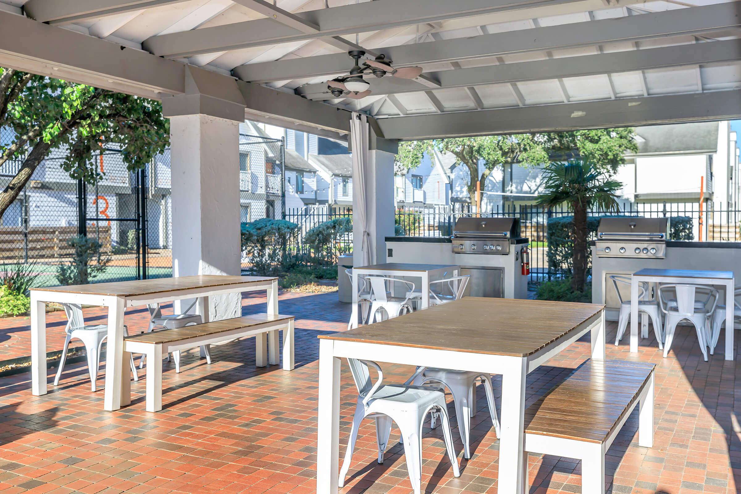 Outdoor communal space with several white picnic-style tables and benches under a covered area. Two stainless steel grills are visible, surrounded by greenery. Fenced area in the background with a glimpse of residential buildings. The flooring consists of red bricks, creating a warm and inviting atmosphere.