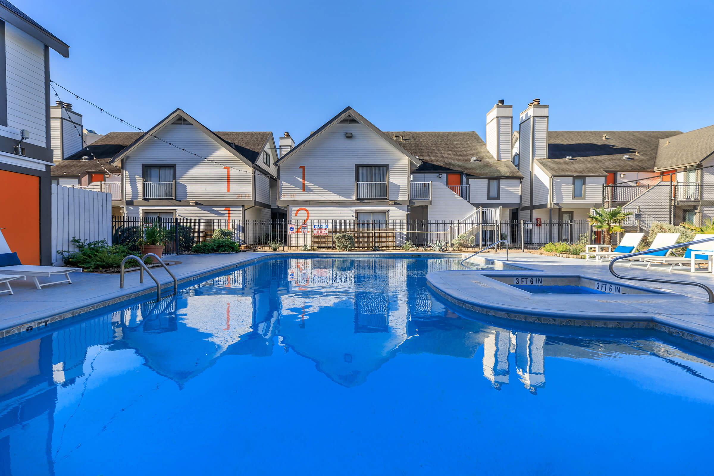 A serene swimming pool surrounded by lounge chairs, with clear blue water reflecting the sky. In the background, several two-story residential buildings with sloped roofs and colorful accents are visible. The area is well-maintained, with some greenery and a gated fence enclosing the pool.