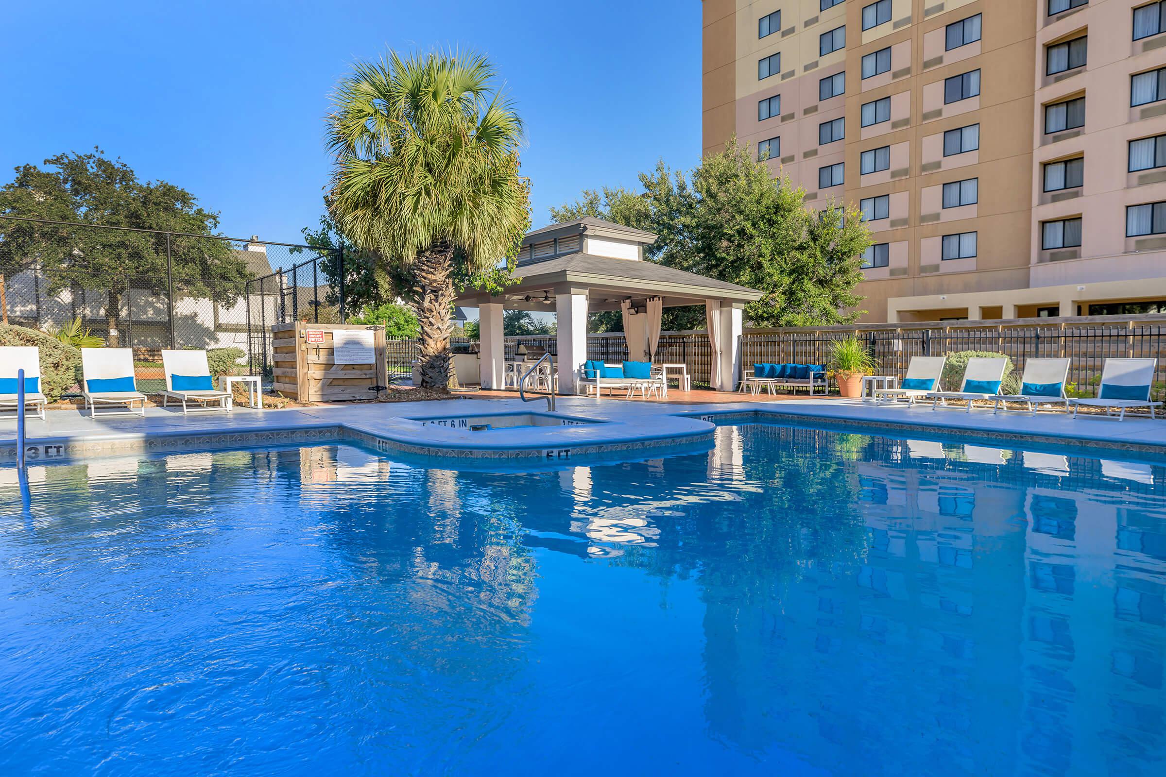 A clear swimming pool surrounded by lounge chairs and palm trees, with a backdrop of a beige hotel building. The sunlight reflects off the water, creating a bright and inviting atmosphere. A covered area nearby provides shade, enhancing the poolside relaxation space.