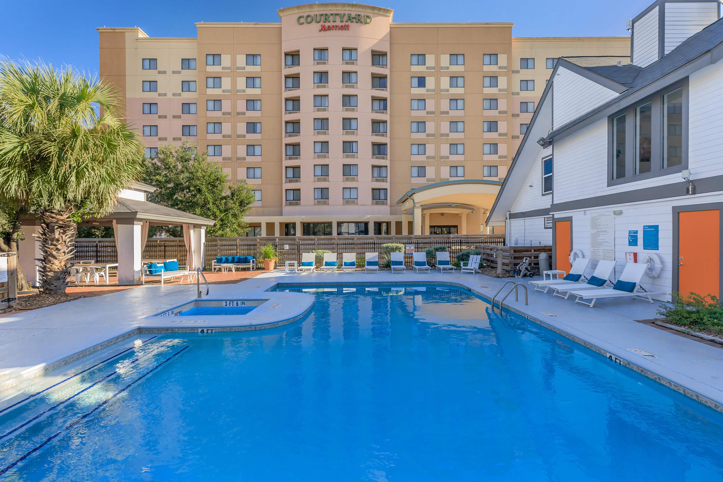 A swimming pool with lounge chairs around it, located in an outdoor area. Behind the pool, there is a hotel building with multiple floors and windows. Sunlight brightens the scene, and palm trees are present, adding to the tropical ambiance. The area appears well-maintained and inviting.