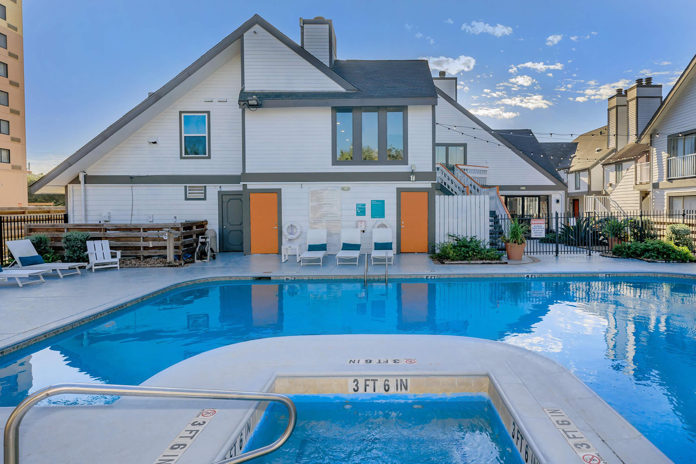 A swimming pool surrounded by lounge chairs, with a spa area in the foreground. A spacious, well-maintained building with orange doors is visible in the background. The scene is set on a clear day, with blue skies and a few clouds, creating a relaxing atmosphere.