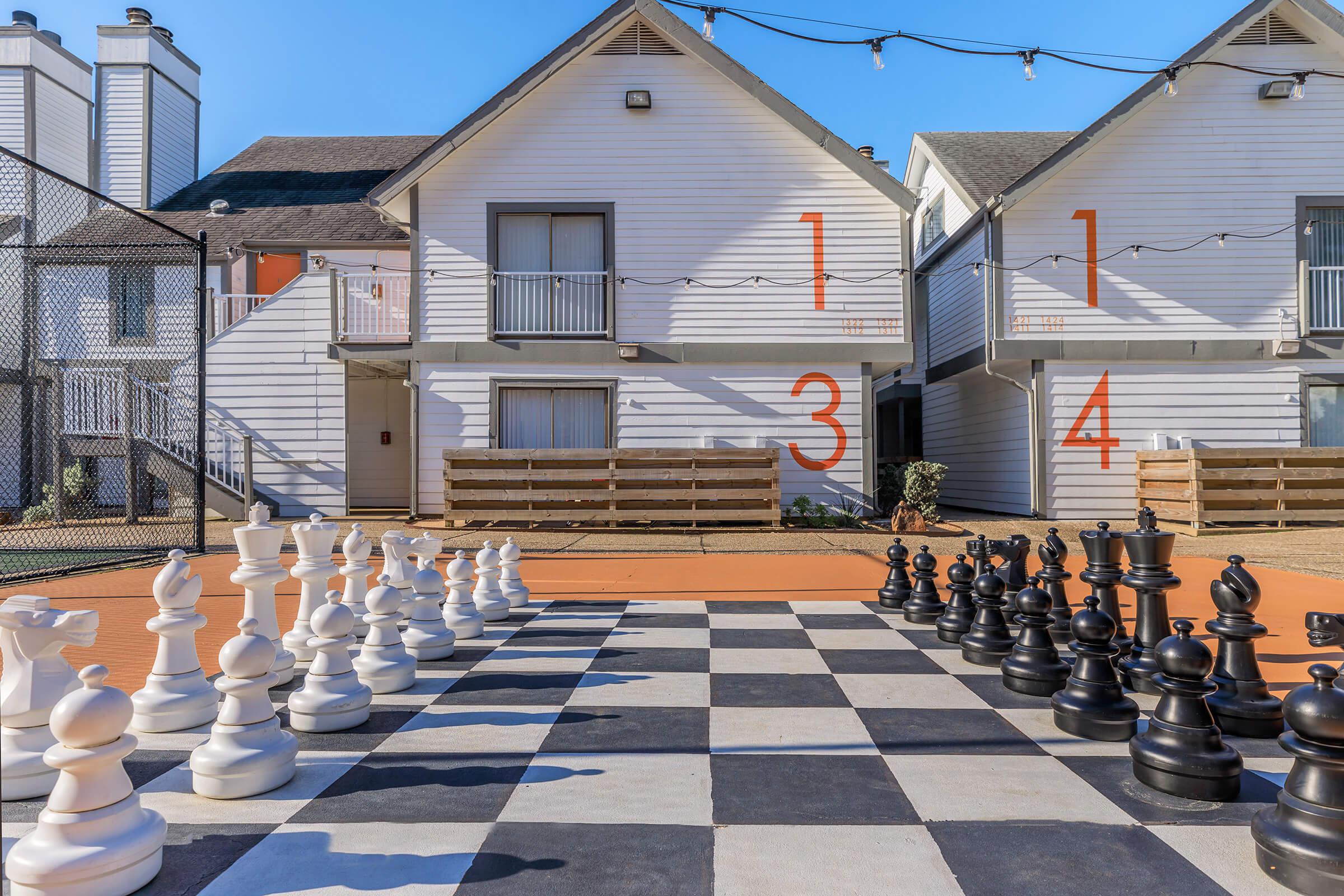 A large outdoor chessboard with white and black oversized chess pieces set up for play. In the background, two white buildings are visible, each with large numbers painted on their walls (1, 3, and 4). The setting is sunny, with a clear blue sky and string lights overhead, creating a vibrant atmosphere.