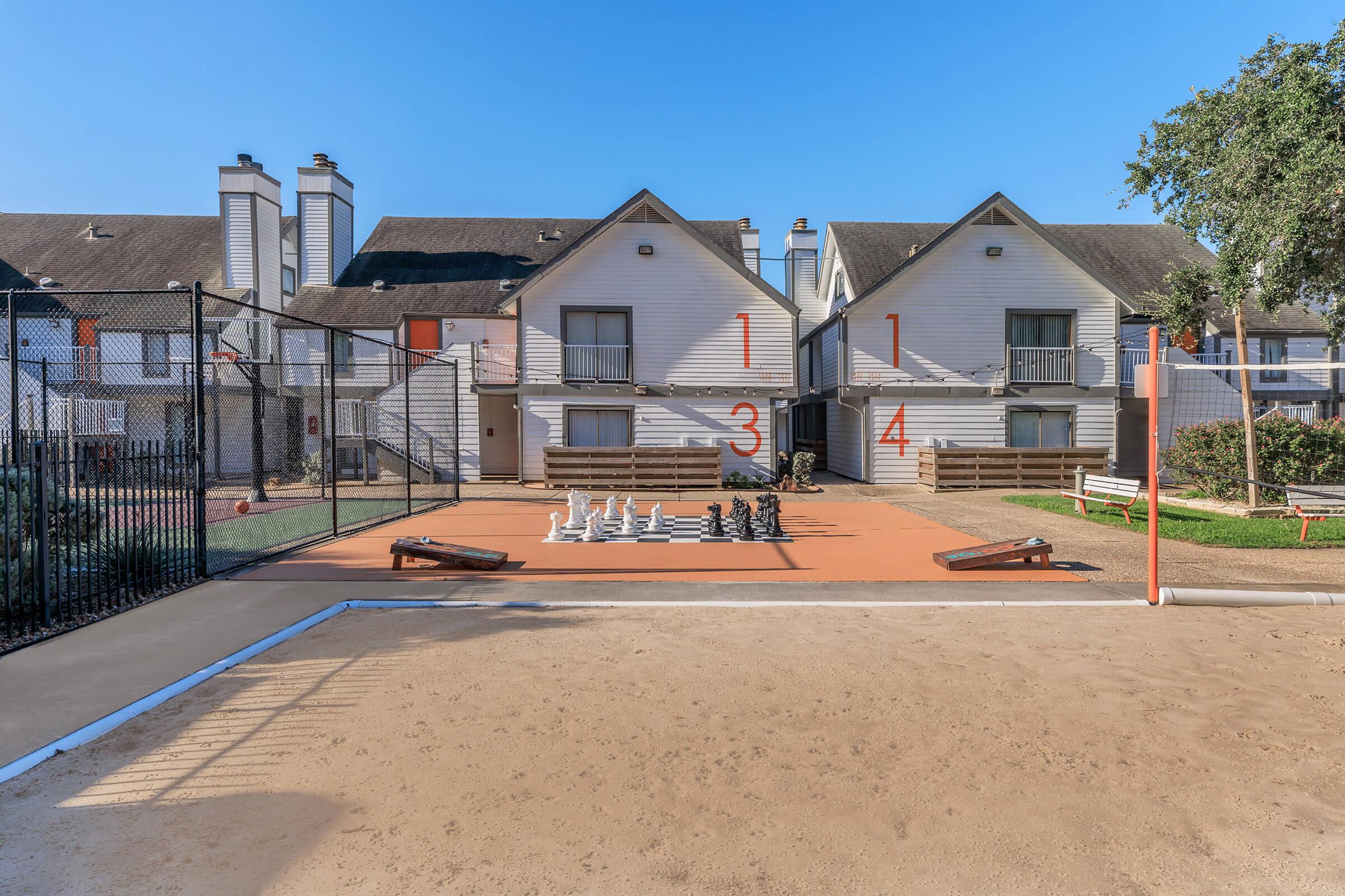 A view of an outdoor recreational area featuring a large chess set on a court, surrounded by two-story residential buildings. The chess pieces are oversized and arranged on an orange surface. In the background, there are benches and a fenced area, with trees providing some shade under a clear blue sky.