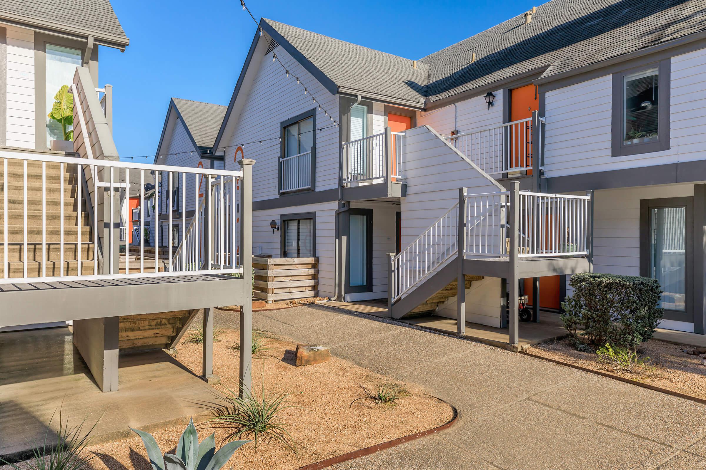 A sunny courtyard view of a multi-unit residential complex featuring two-story buildings with stairs leading to upper units. The exterior is composed of light colors, and there are small shrubs and decorative plants in the landscape, creating a welcoming atmosphere.