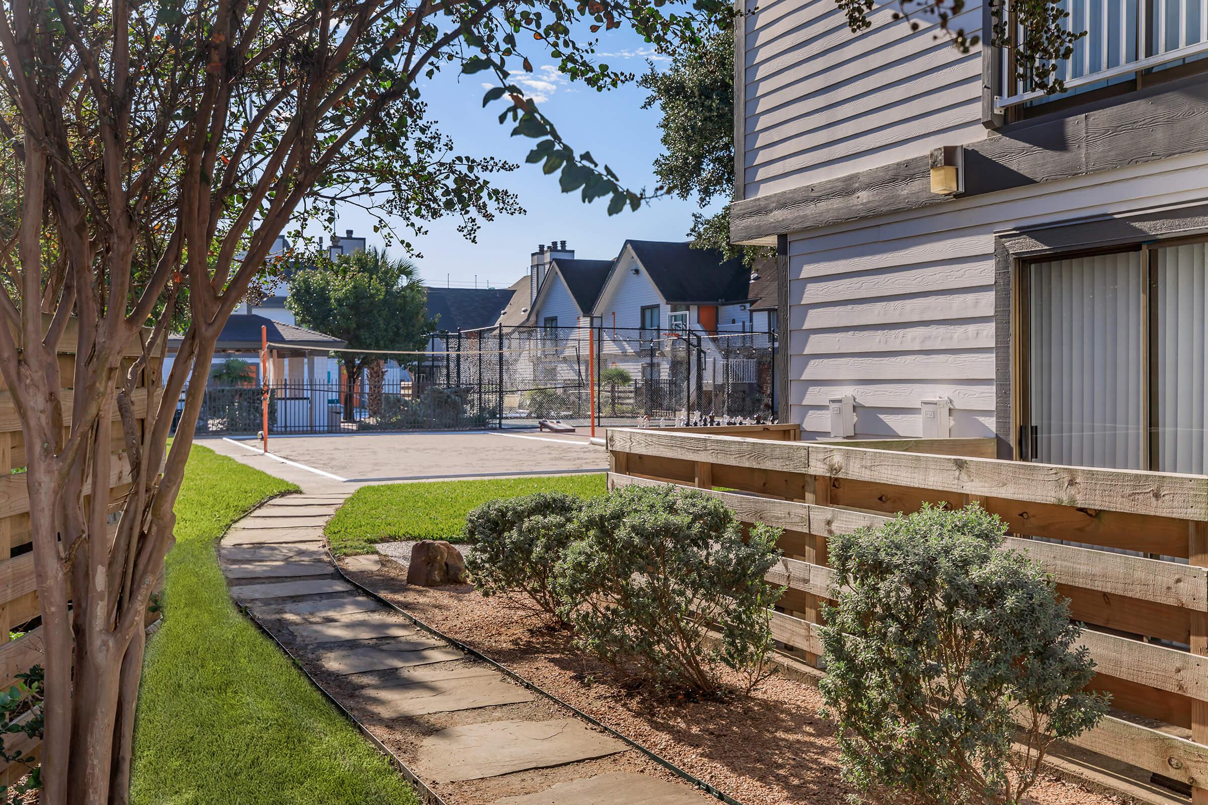 A view of a landscaped pathway leading to a fenced outdoor area, with a mix of grass and gravel. On the left, there are low bushes and a wooden fence, while the background features a mix of residential buildings and a sports court. The scene is bright and sunny, conveying a peaceful atmosphere.