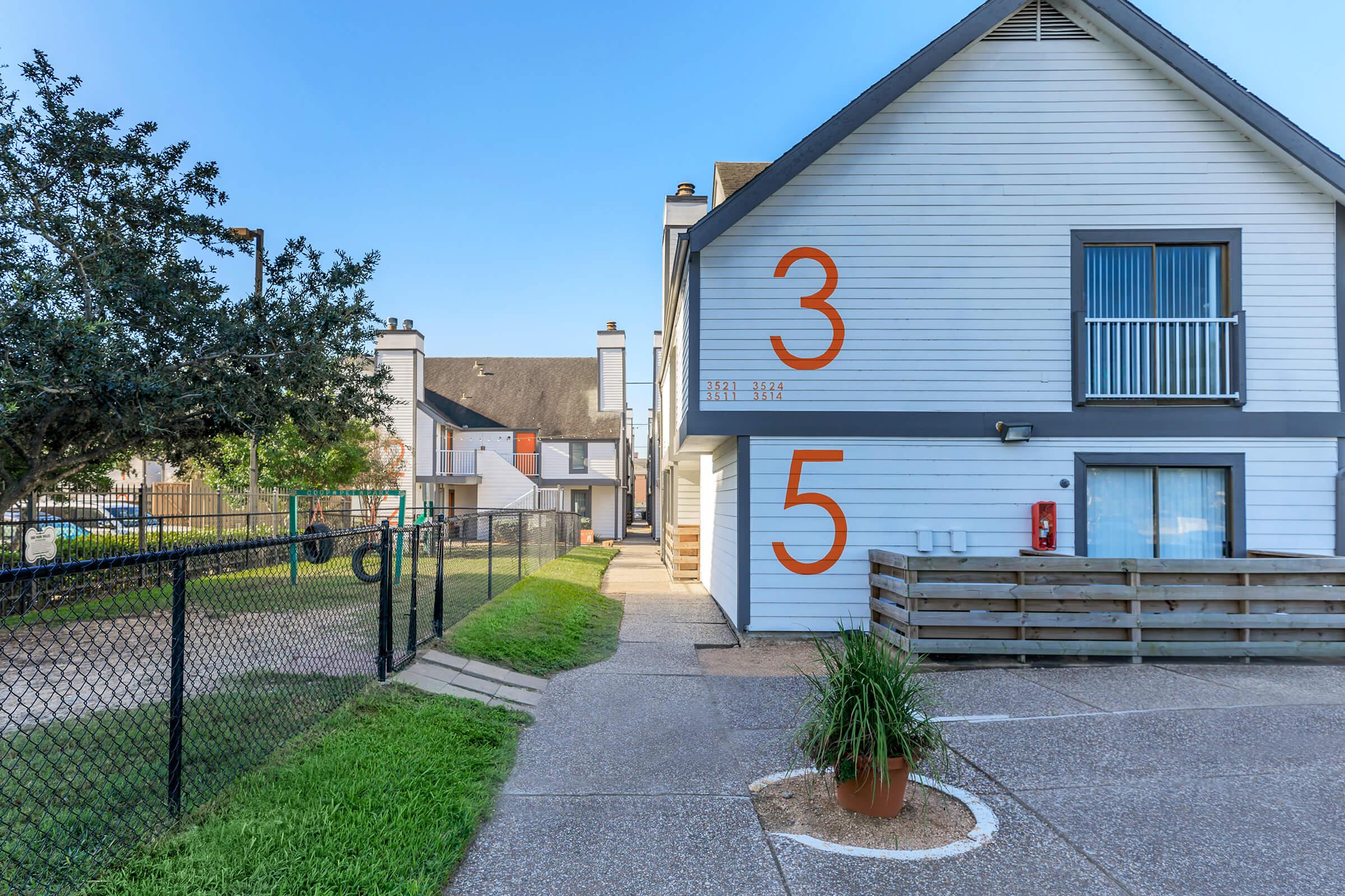 A two-story building with large orange numbers "3" and "5" on the exterior wall. A pathway leads to the entrance, surrounded by greenery and a fenced area. There's a fire extinguisher mounted on the wall, and the sky is clear. The setting appears to be a residential or apartment community.