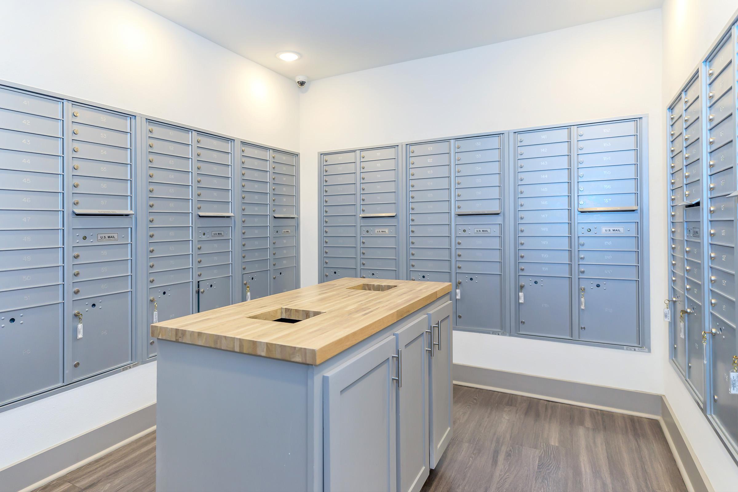 A modern mail room featuring multiple rows of silver mailboxes mounted on the walls. In the center, there is a wooden countertop with an opening, providing a space for package drop-off or pick-up. The walls are painted white, and the flooring is a dark wood laminate. A ceiling light illuminates the area.