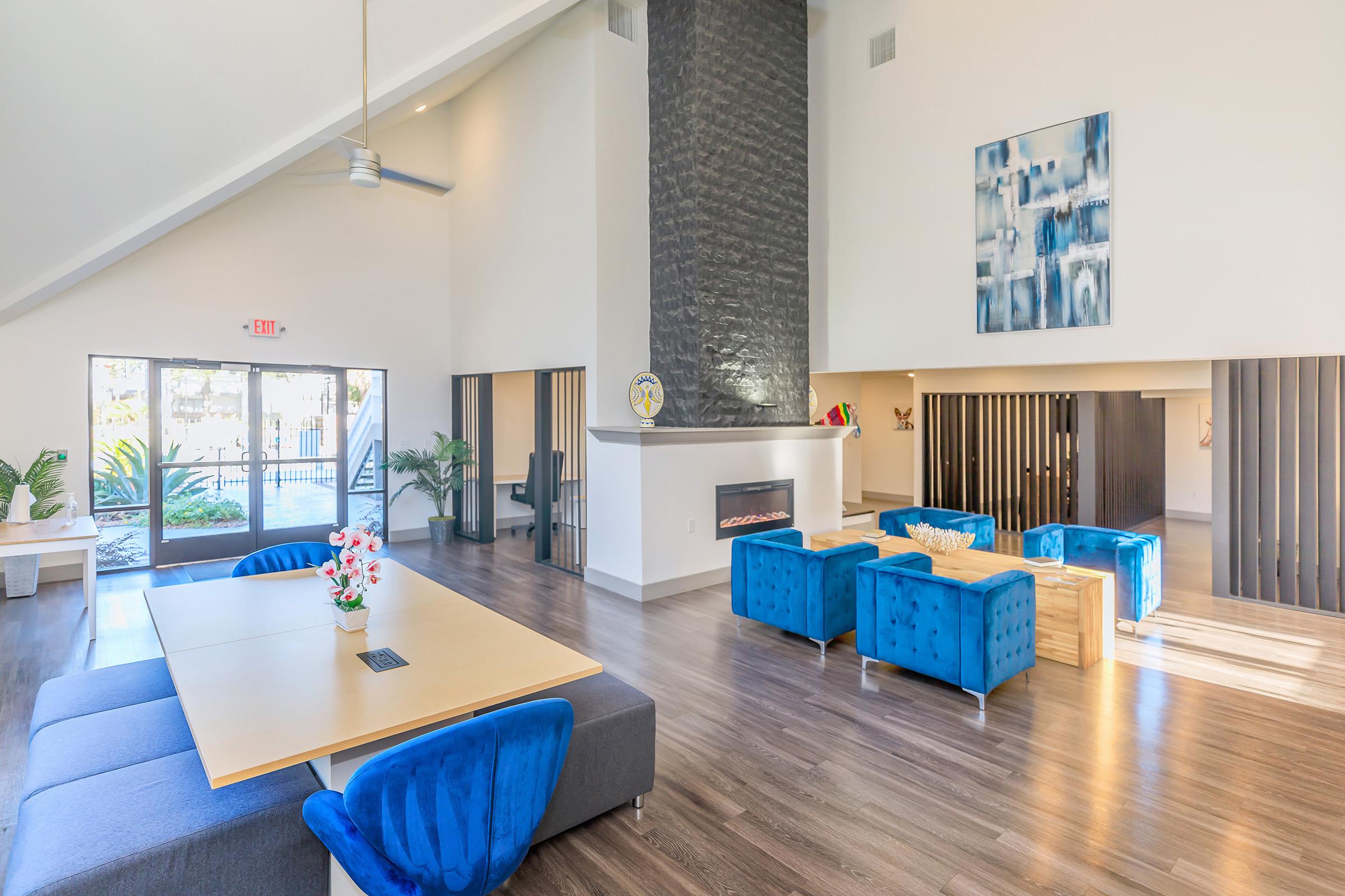 A spacious, modern lounge area featuring a large table surrounded by blue upholstered chairs, a cozy fireplace, and large windows letting in natural light. Decorative elements include a floral arrangement on the table and artwork on the wall, creating a welcoming atmosphere.