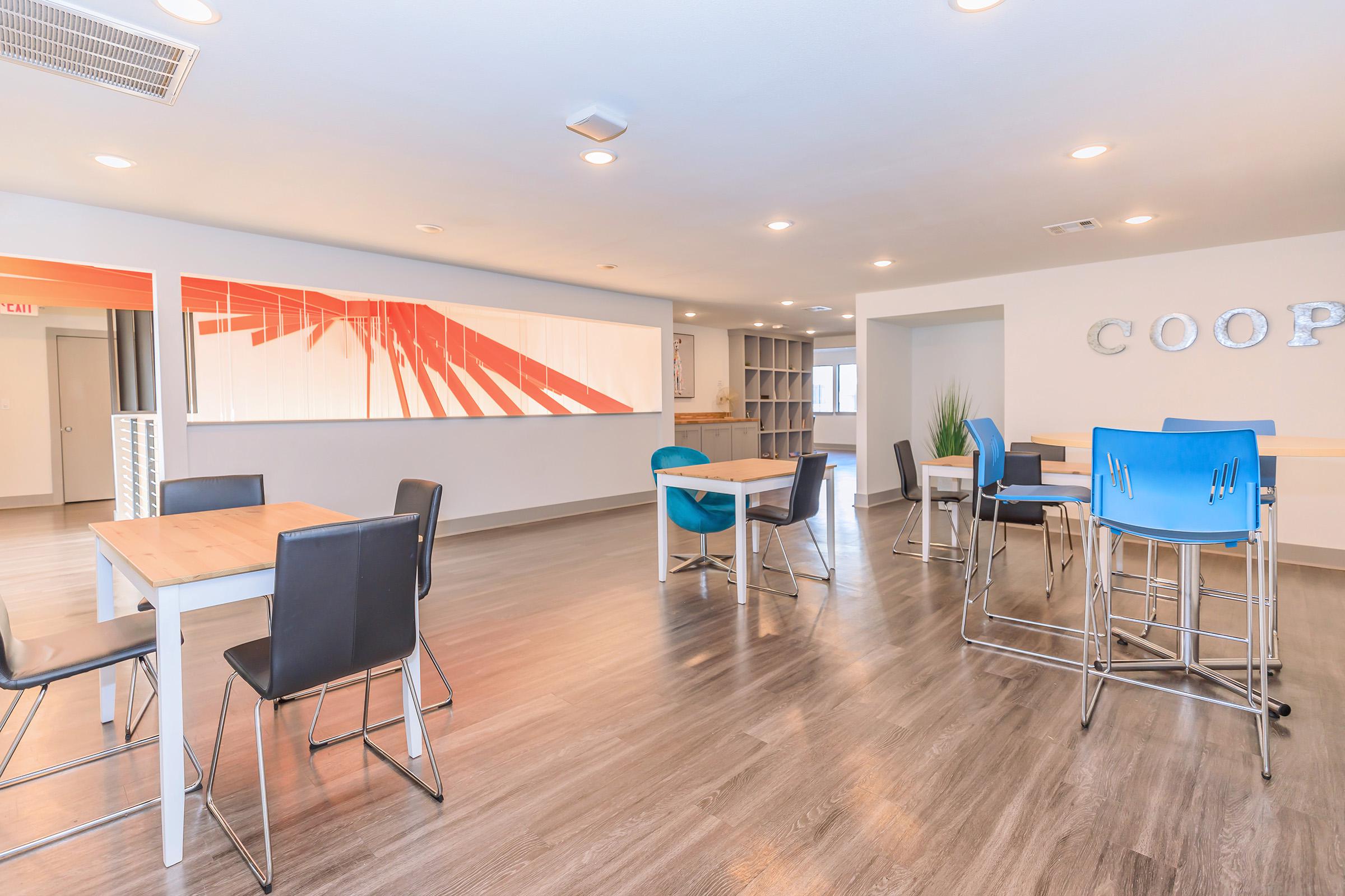 A spacious and modern communal area featuring light wood tables with black and blue chairs. The interior has a neutral color palette with orange accents on the wall art, which displays diagonal lines. The flooring is a sleek gray, contributing to a clean and inviting atmosphere.