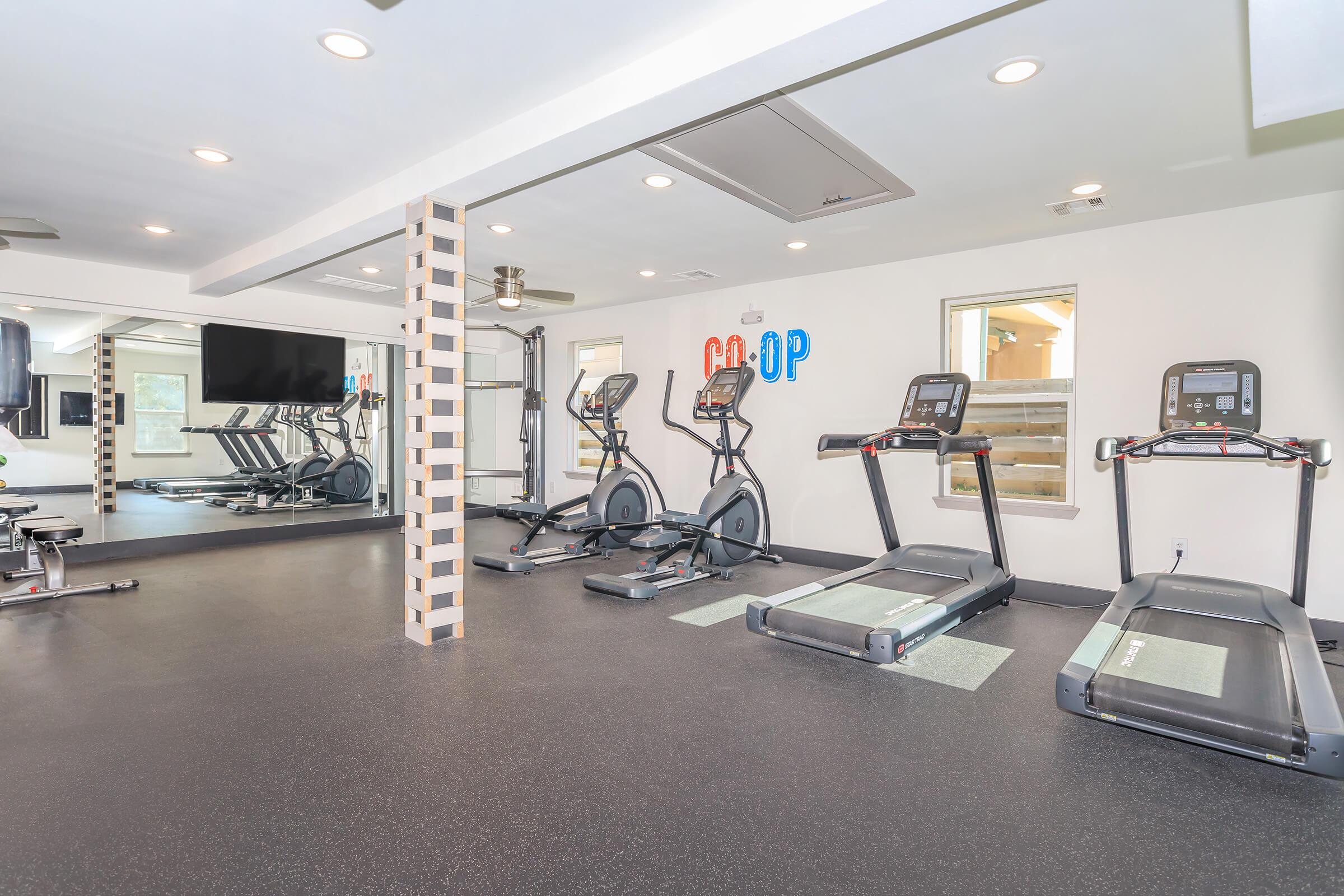 A modern gym interior featuring various fitness equipment, including two treadmills and an elliptical machine. The space is well-lit with a mirror on one wall, a flat-screen TV, and colorful wall decor spelling "CO-OP." The floor is covered in black rubber mats for safety and comfort.