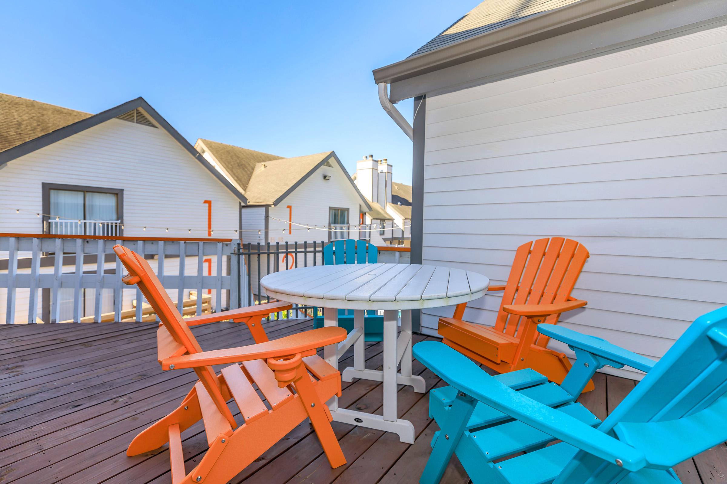 A wooden deck features a round white table surrounded by brightly colored adirondack chairs in orange and turquoise. The setting includes two rooftops in the background under a clear blue sky.