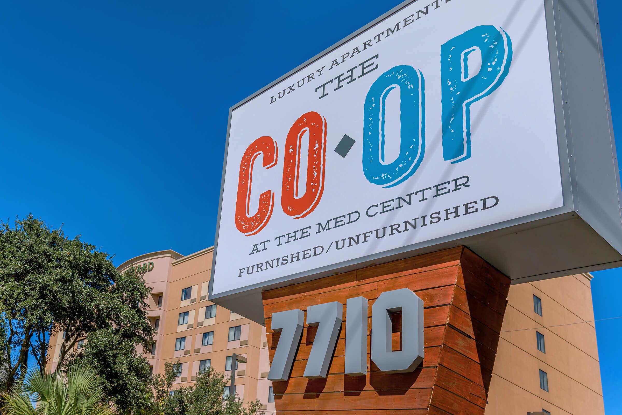 Sign for "The Co-Op," a luxury apartment community located at the Med Center. The sign highlights it offers furnished and unfurnished units, with the address "7710" prominently displayed. Bright blue sky in the background complements the colorful design of the sign.